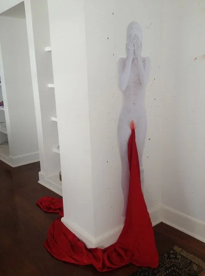 A sculpture of a woman with long hair, covering her face with hands, and a red cloth hanging from the sculpture's waist to the floor.