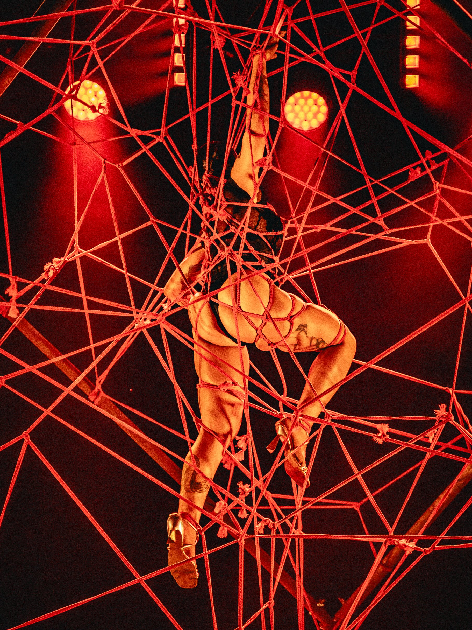 A performer on aerial silk hangs upside down amidst a red web of ropes with stage lighting in the background.