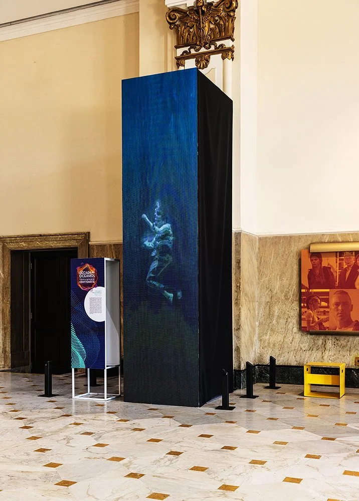 An art installation in a museum featuring a tall, black rectangular display with a projected image of a person in mid-air, possibly performing a dance or acrobatics, surrounded by informational signage.