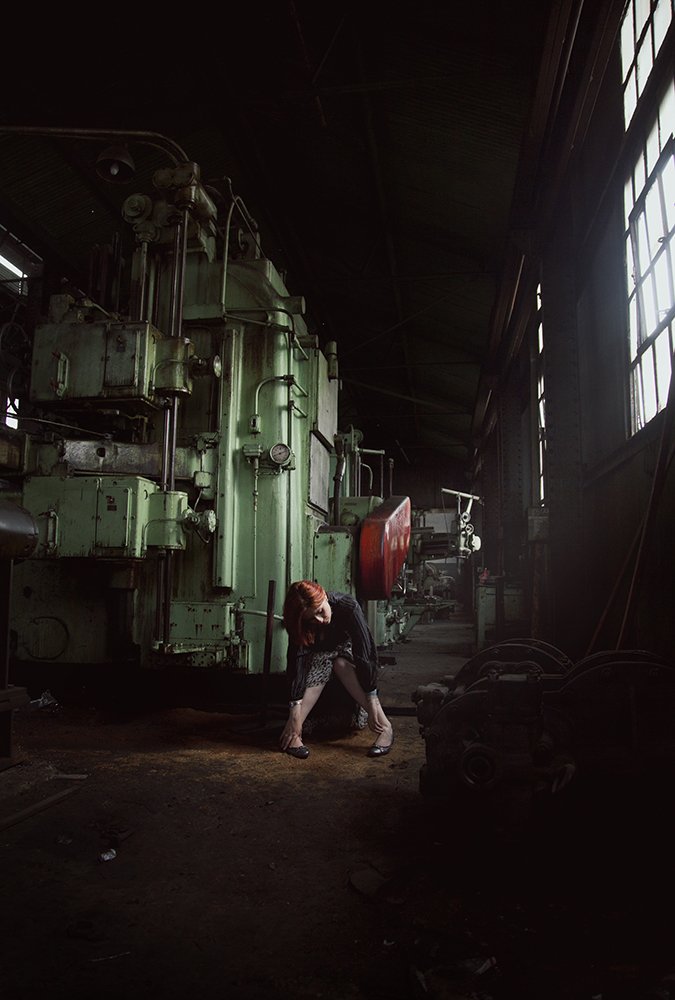 A woman crouching and leaning forward inside an industrial workshop surrounded by large, green machinery. The workshop is dimly lit with natural light coming through windows on the right.