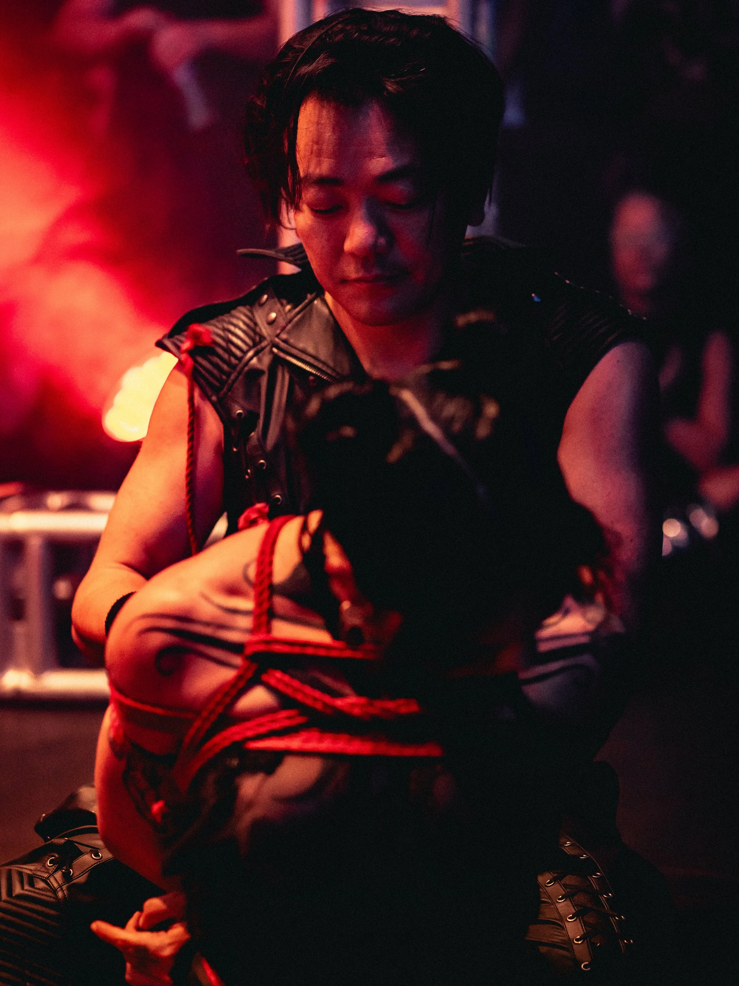 Close-up of a person with short black hair wearing a leather vest, engaged in a dramatic scene with another person. The background has red and purple lighting, suggesting a tense or intense atmosphere.