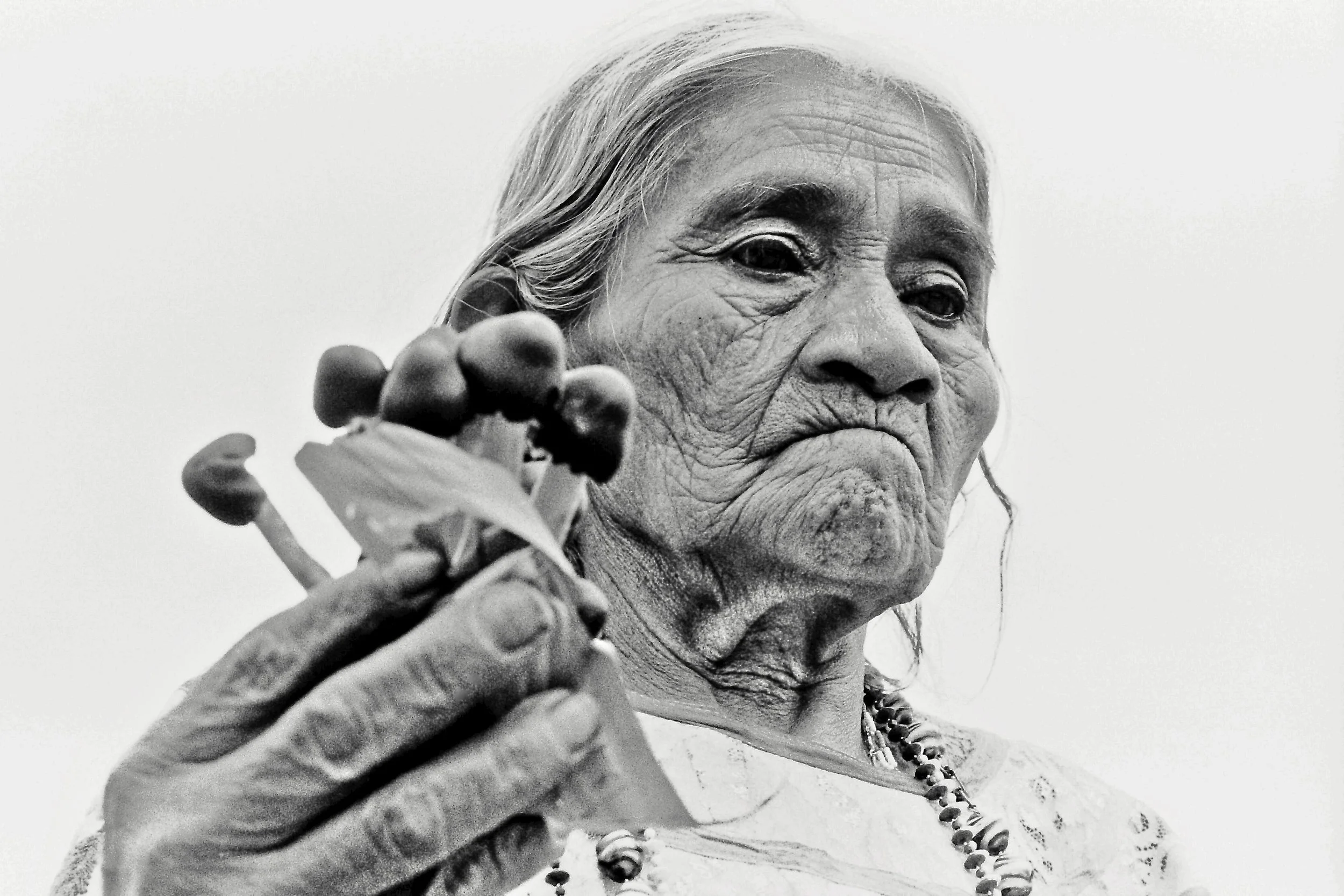 A black and white close-up photo of an elderly woman with long hair, wearing a beaded necklace, holding a cluster of grapes and a leaf in her hand, with a serious expression.