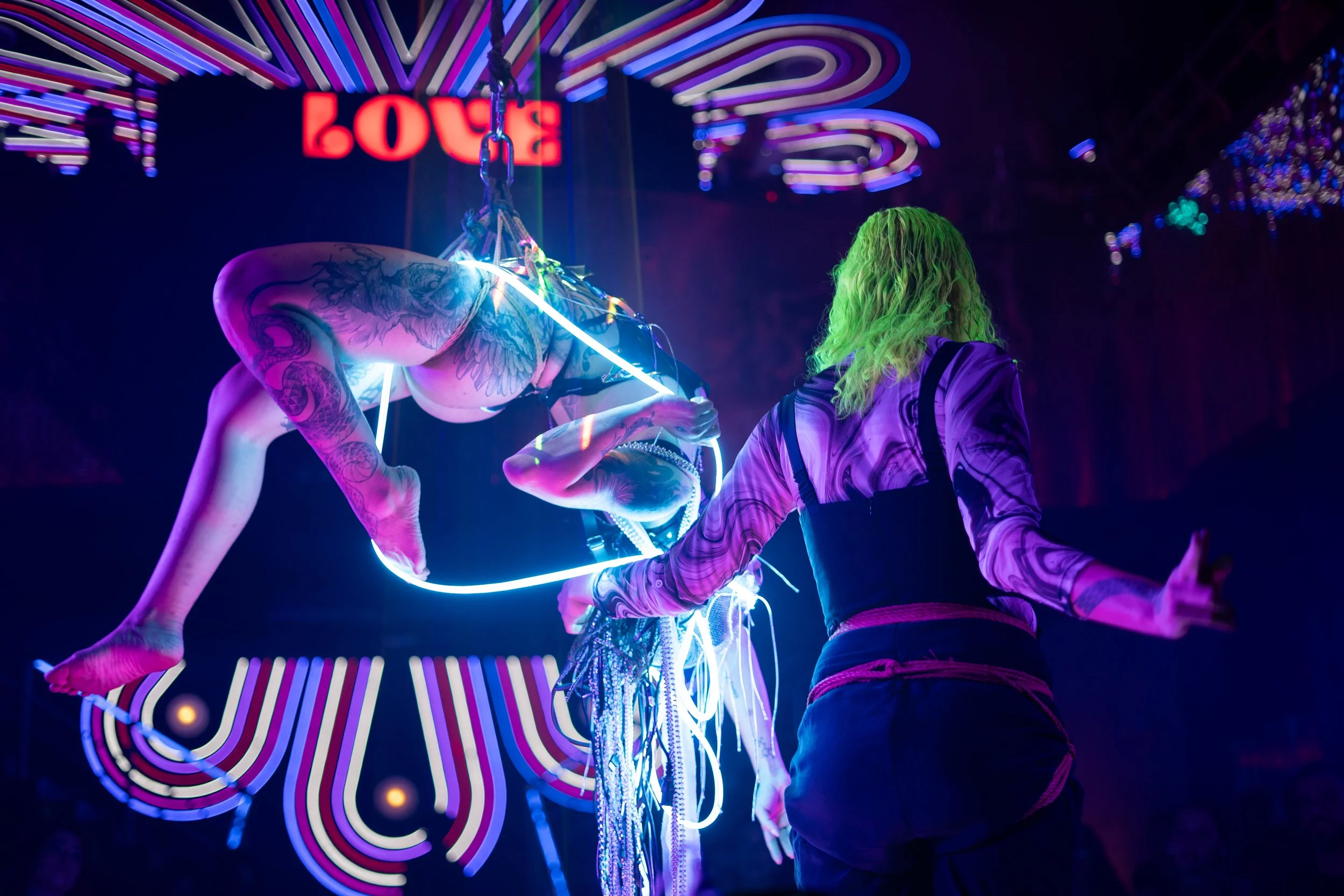 A performer in a black outfit with tattoos on their arm, hanging upside down on a neon-lit trapeze, illuminated by purple, blue, and pink lights, while a person with green hair and a purple patterned shirt watches.