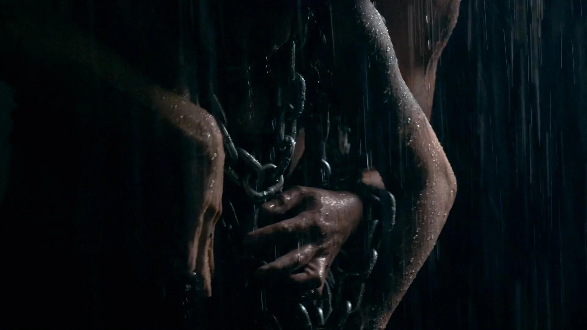 A person with wet skin holding a large chain, with rain falling around them in a dark setting.