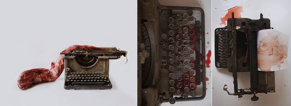 An old typewriter with a red cloth or piece of fabric draped behind it, and the typewriter is damaged and missing parts, placed next to a box of empty typewriter ribbon spools and a small mechanical device on a white surface with bloodstains.