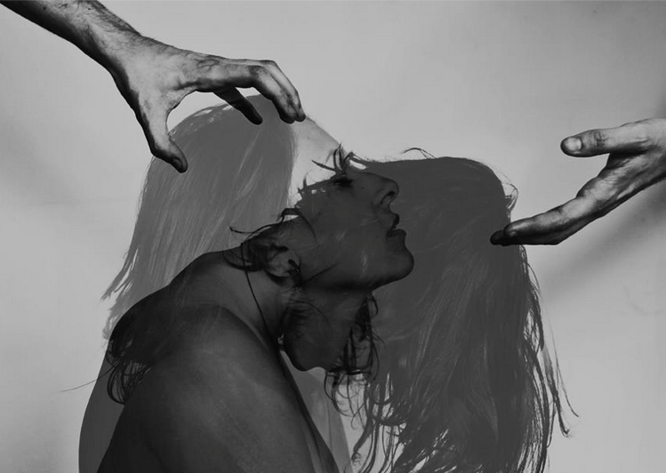 Black and white photo of a woman with long hair, her silhouette visible through her hair, with two hands reaching toward her head from opposite sides.