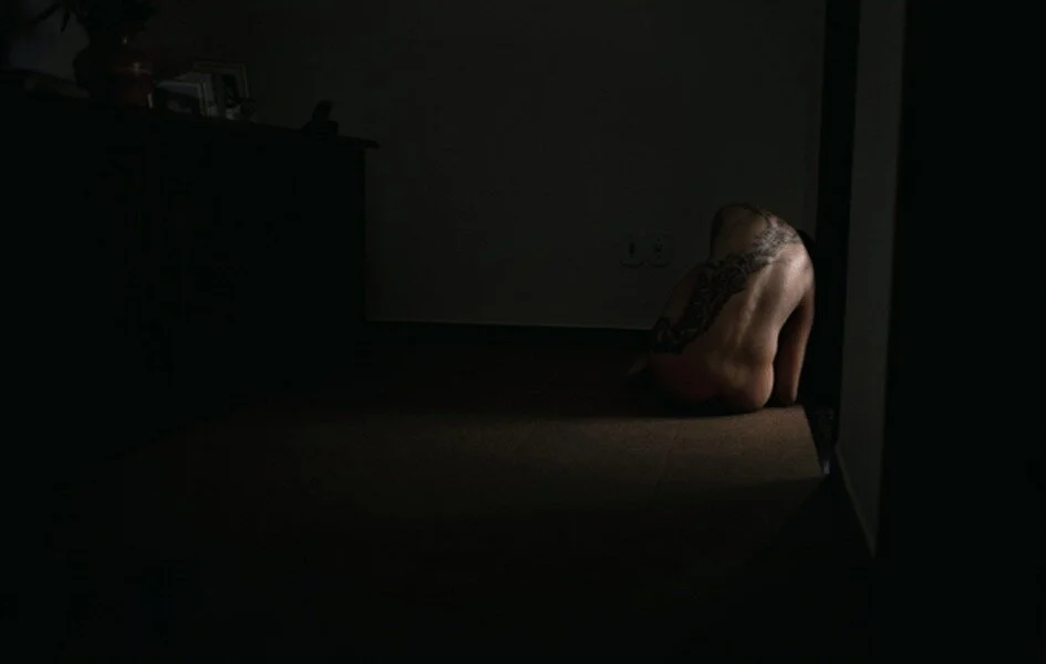 A person with a tattoo on their back sitting on the floor in a dark room facing the corner.