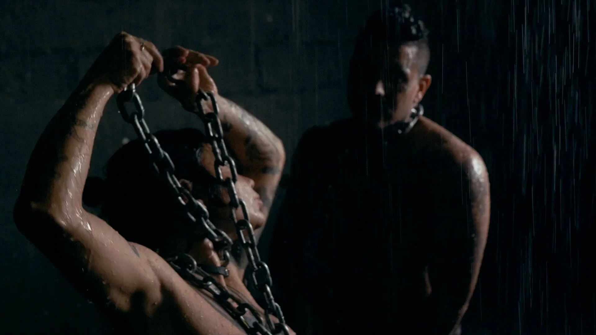 Two shirtless men with tattoos, chained together, standing in the rain at night, one with his eyes closed, the other looking at him in the dark.
