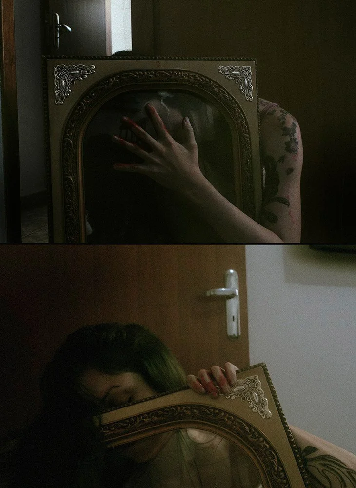 A person with tattoos holding a mirror in front of their face, with a hand reaching into the mirror from behind. The mirror has an ornate gold frame, and the scene is in a dimly lit room with a wooden door and a white door handle in the background.