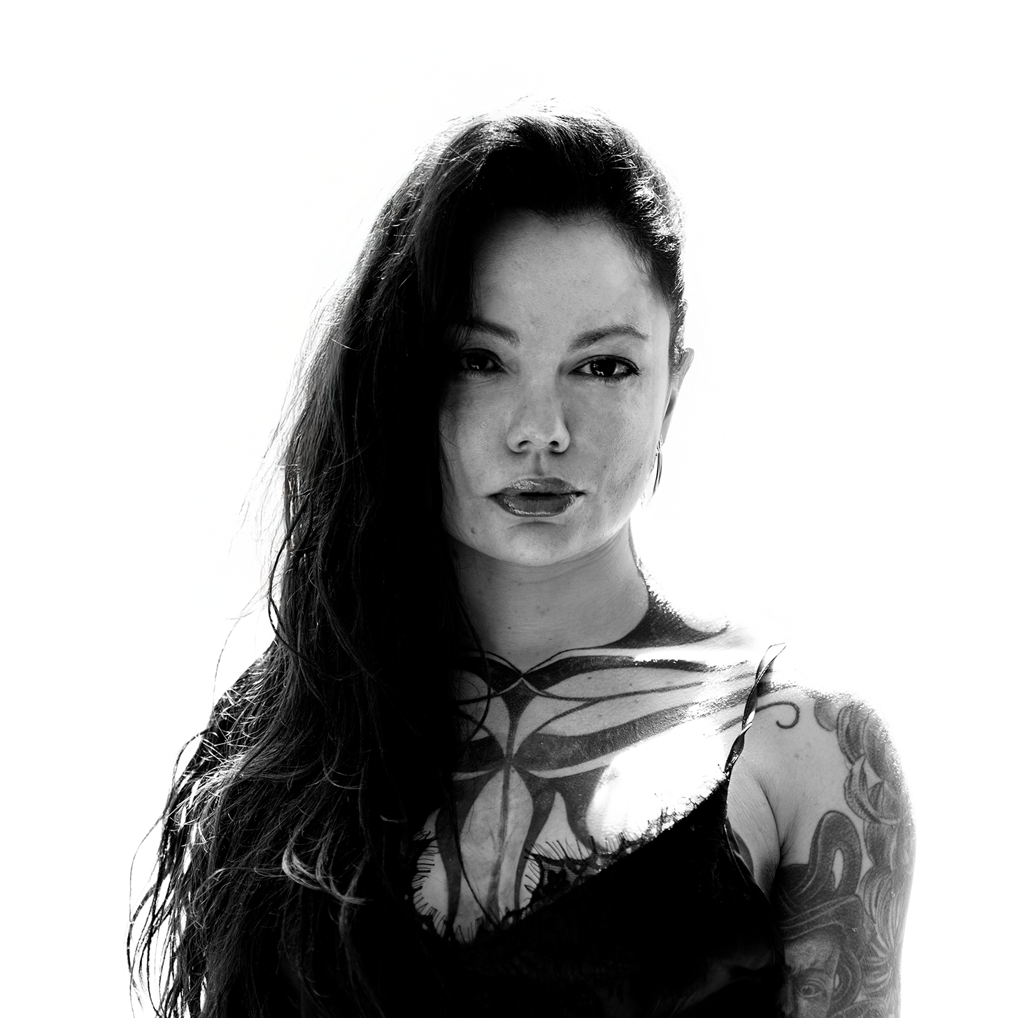 Black and white portrait of a woman with long dark hair, tattoos on her chest and arms, wearing a black top, with a serious expression, on a plain white background.