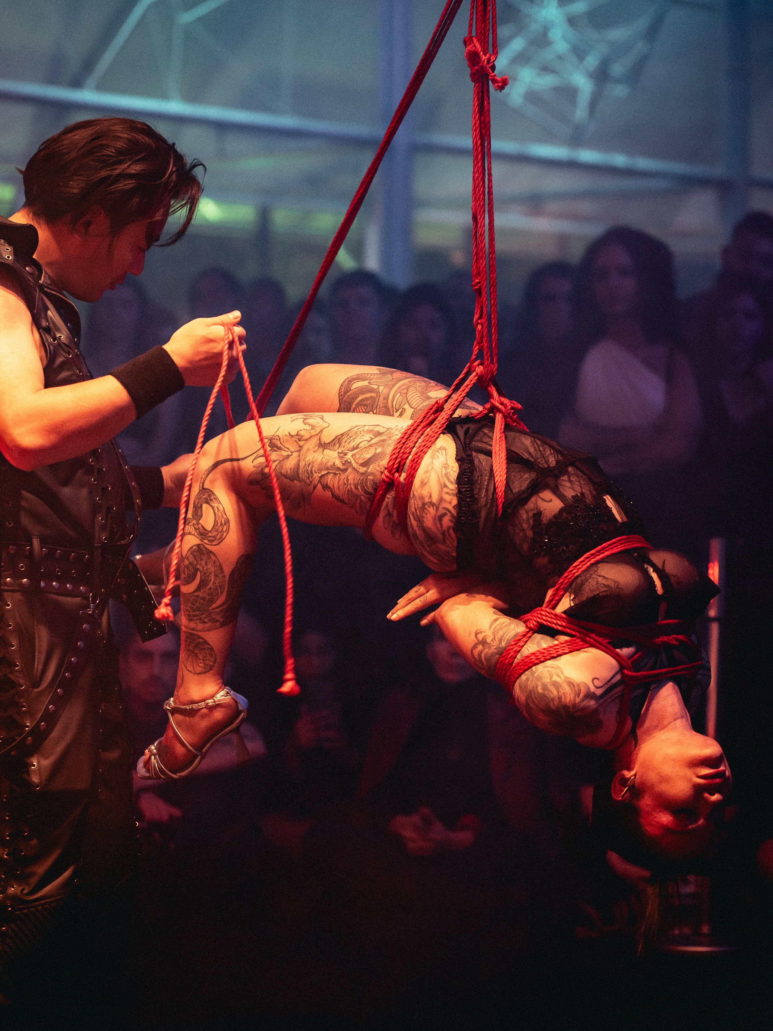 A tattooed woman suspended upside down in a BDSM performance, tied with red ropes, while a performer in leather attire tends to her in front of an audience in dim lighting.
