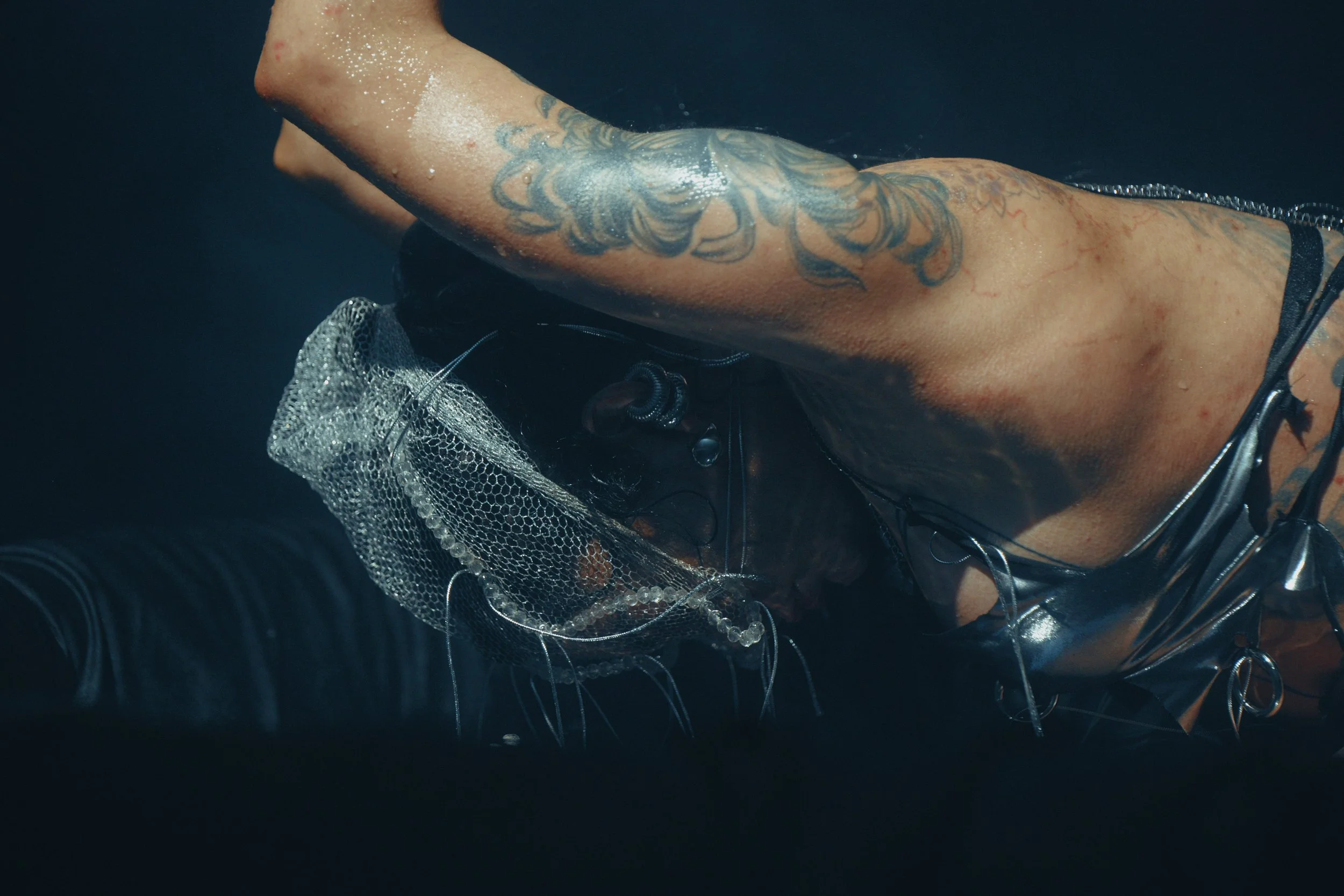 A person with tattoos wearing a black leather outfit and a mesh sleeve, lying down in water with bubbles.