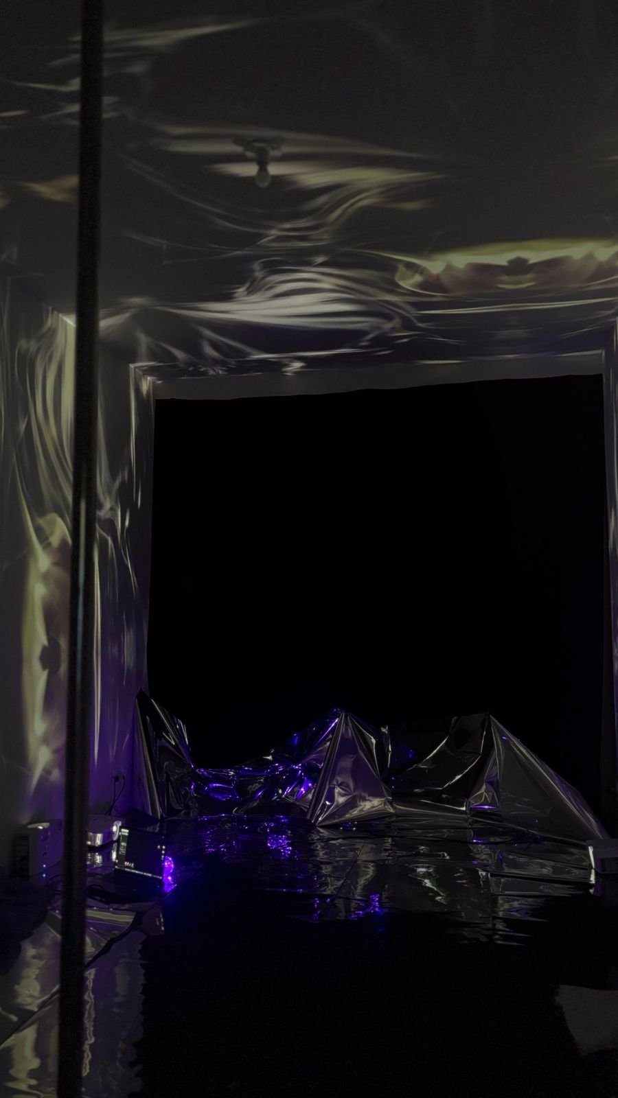 Dark room with reflective metallic crumpled foil surfaces on the floor and walls, illuminated by purple and yellow lights, creating a futuristic, abstract space.