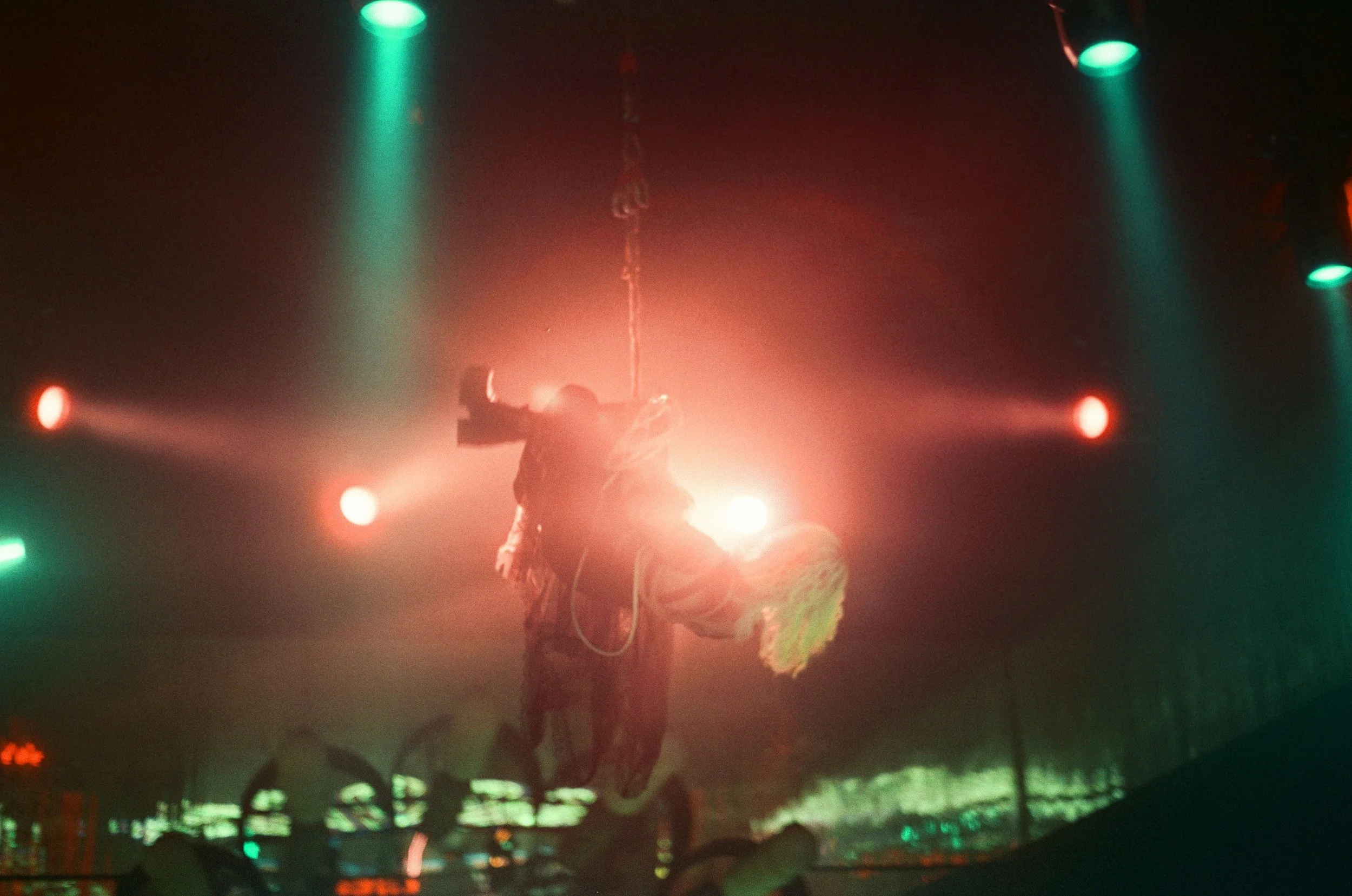A performer hanging from a rope illuminated by stage lights in a dark venue, with colorful spotlights and smoke effects.