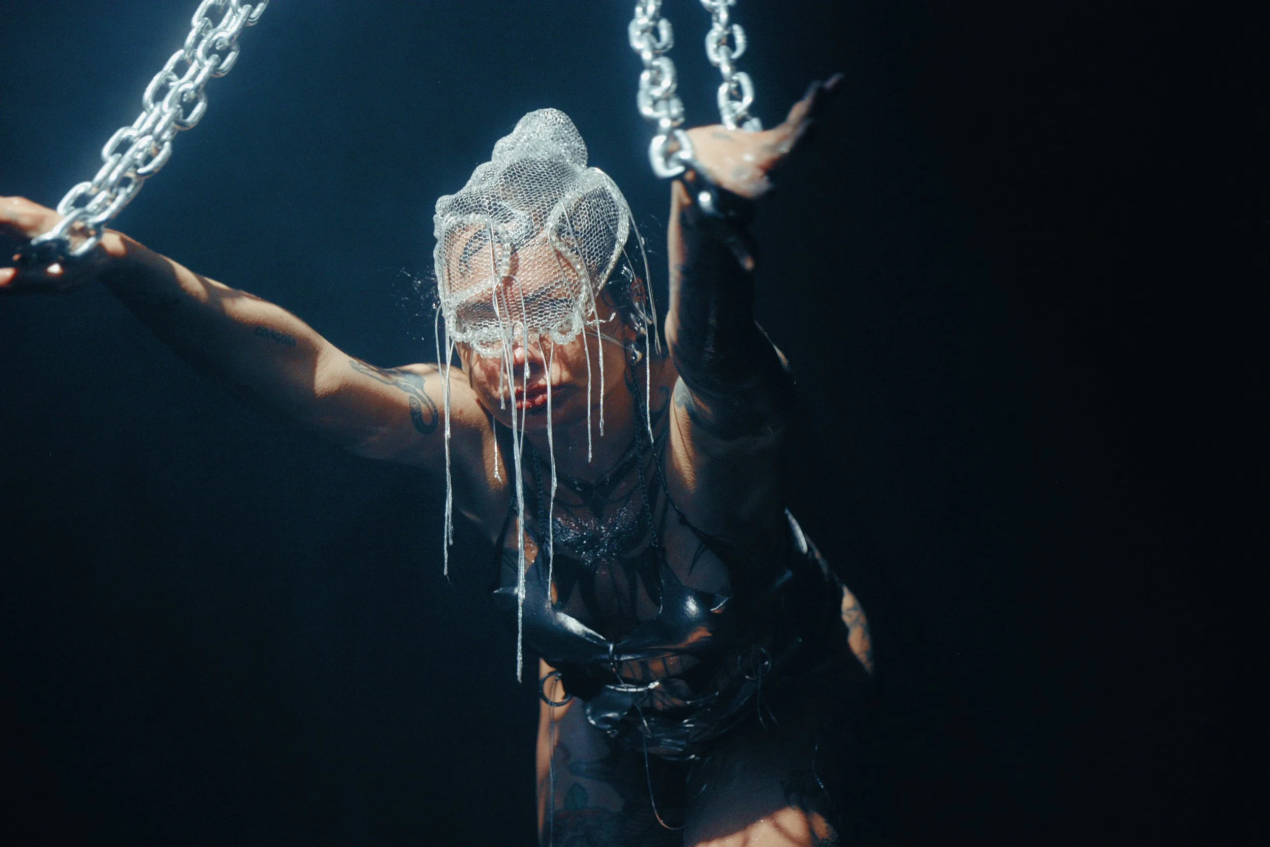 A person with tattoos wearing a black outfit, holding chains, with a netted mask over their face, against a dark background.