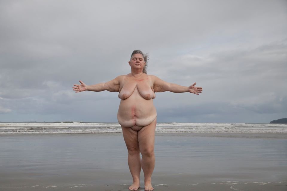 A woman standing with arms outstretched on a beach, facing the camera, with cloudy sky and ocean in the background.