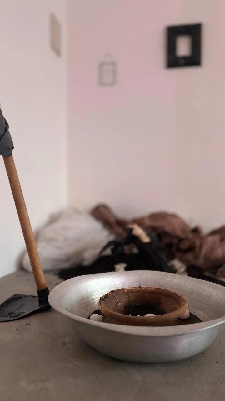 A metal container with a smaller clay bowl inside, some ashes and burnt residue, a shovel with a wooden handle leaning against the container, and some indistinct bedding or cloth in the background.
