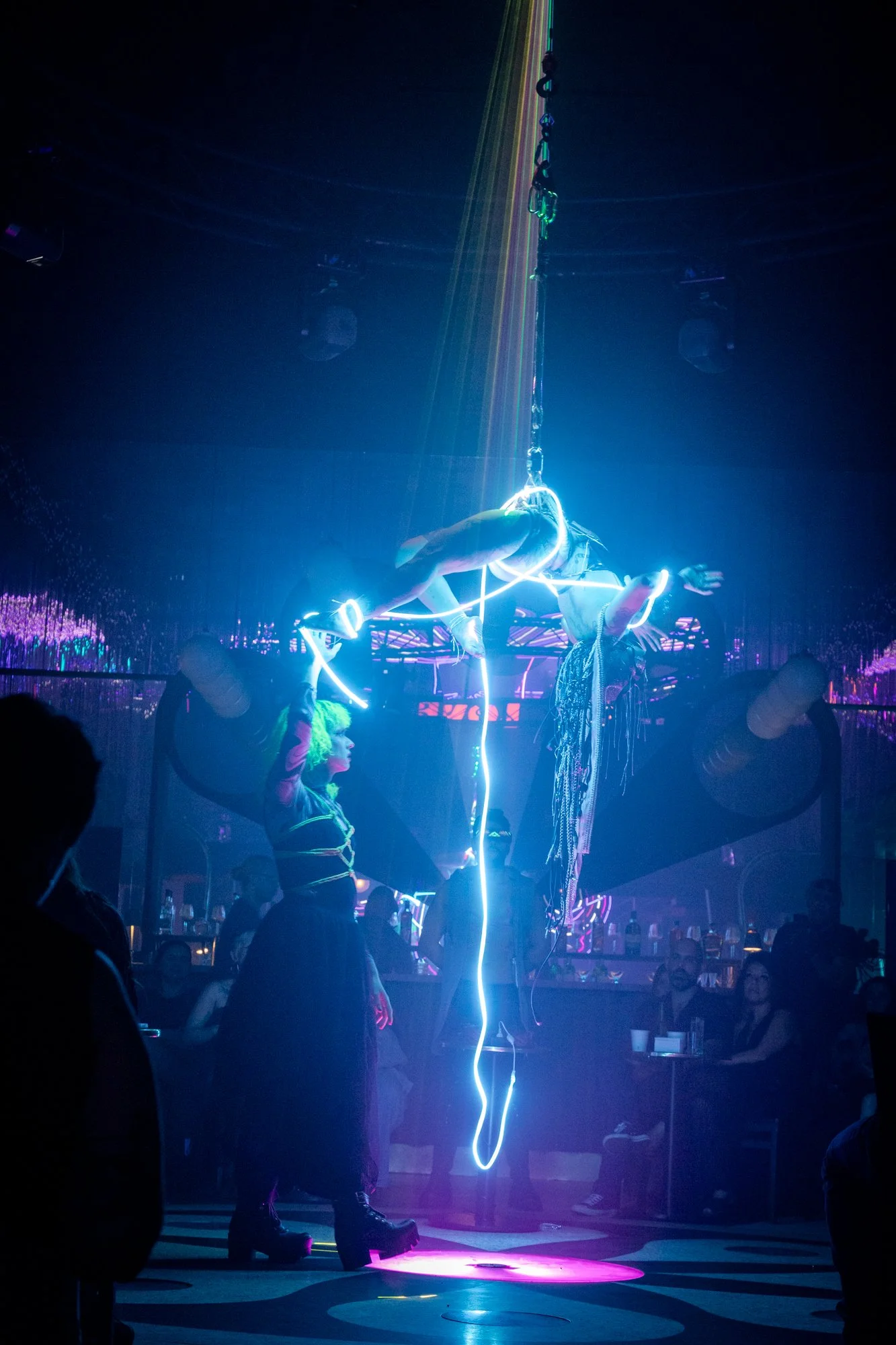 Aerial performers illuminated with neon lights perform an acrobatic act on a stage with a dark background and an audience watching.