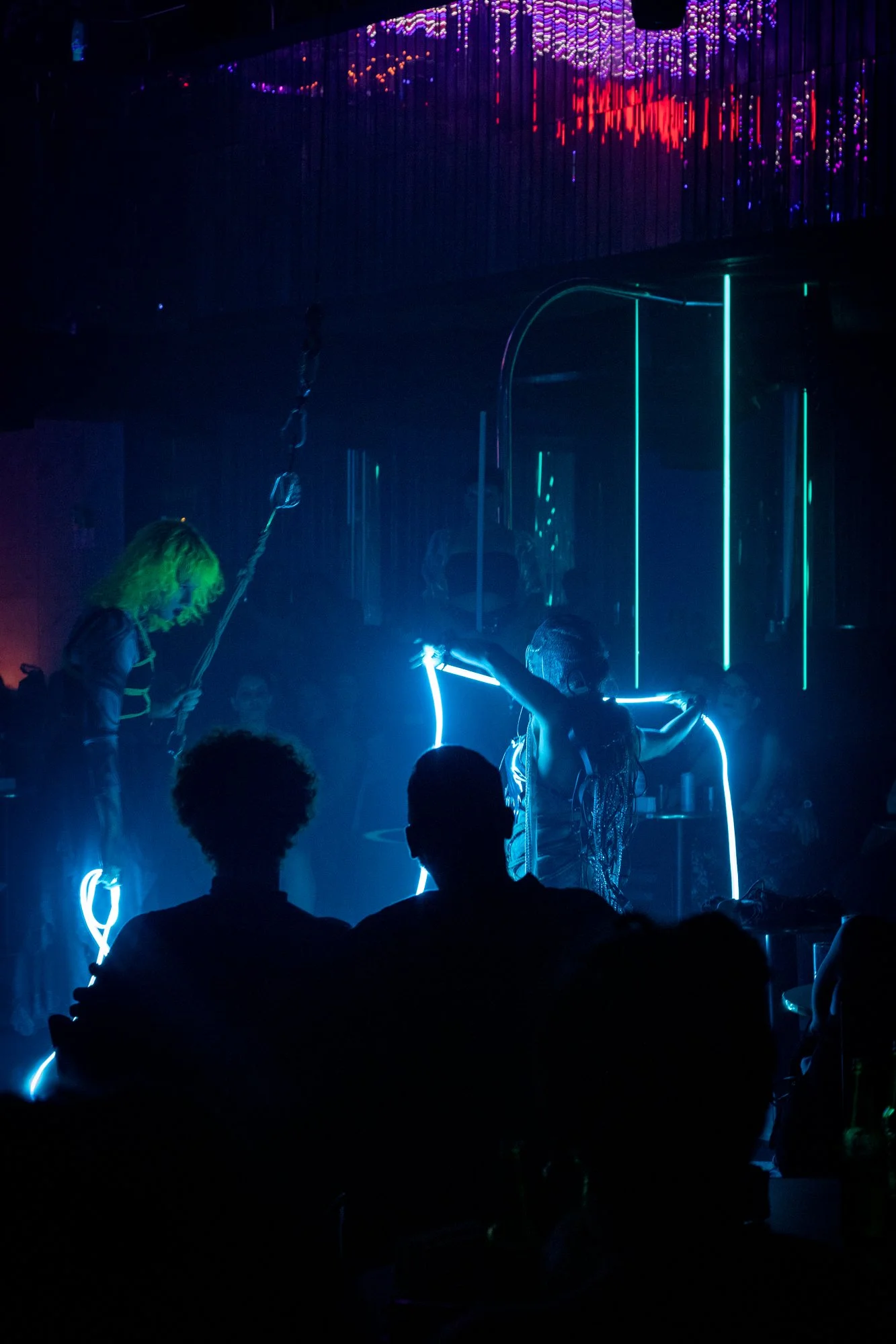Two performers on stage with neon lighting and an audience in a dark club, one holding a chain, the other with glowing accessories.