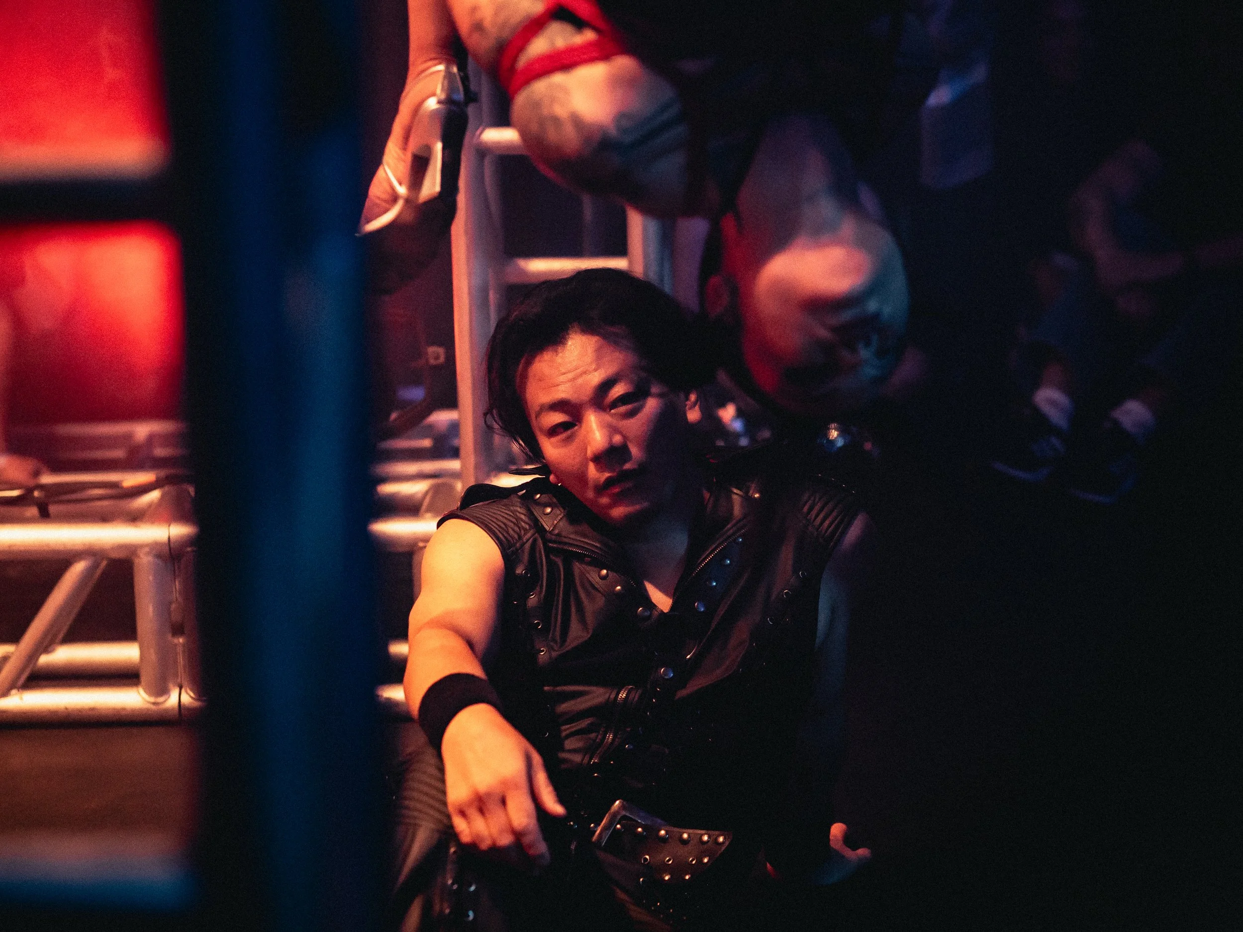 A person with dark hair and a studded black leather outfit sitting on the floor in a dark environment, with another person leaning over them and a figure in the background wearing glasses and black clothing, illuminated by reddish lighting.