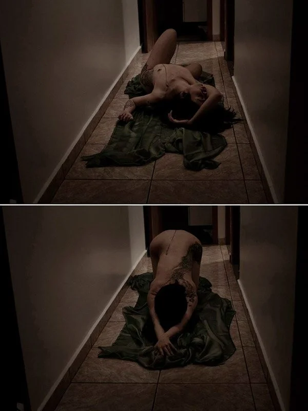 A person lying on a blanket in a narrow hallway, appearing to be undressed and reclining, with a tattoo visible on their back.