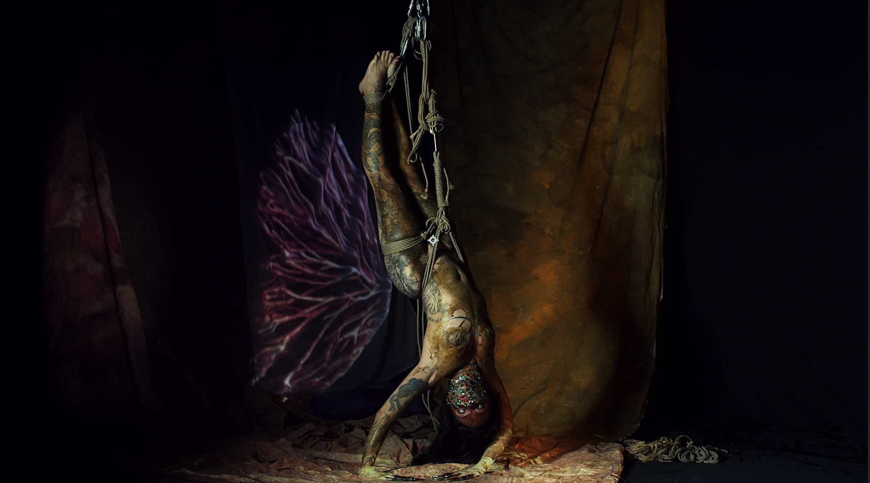 A person with tattoos and face paint performing a contortion pose, hanging upside down with legs tied and suspended by ropes in a dark setting.