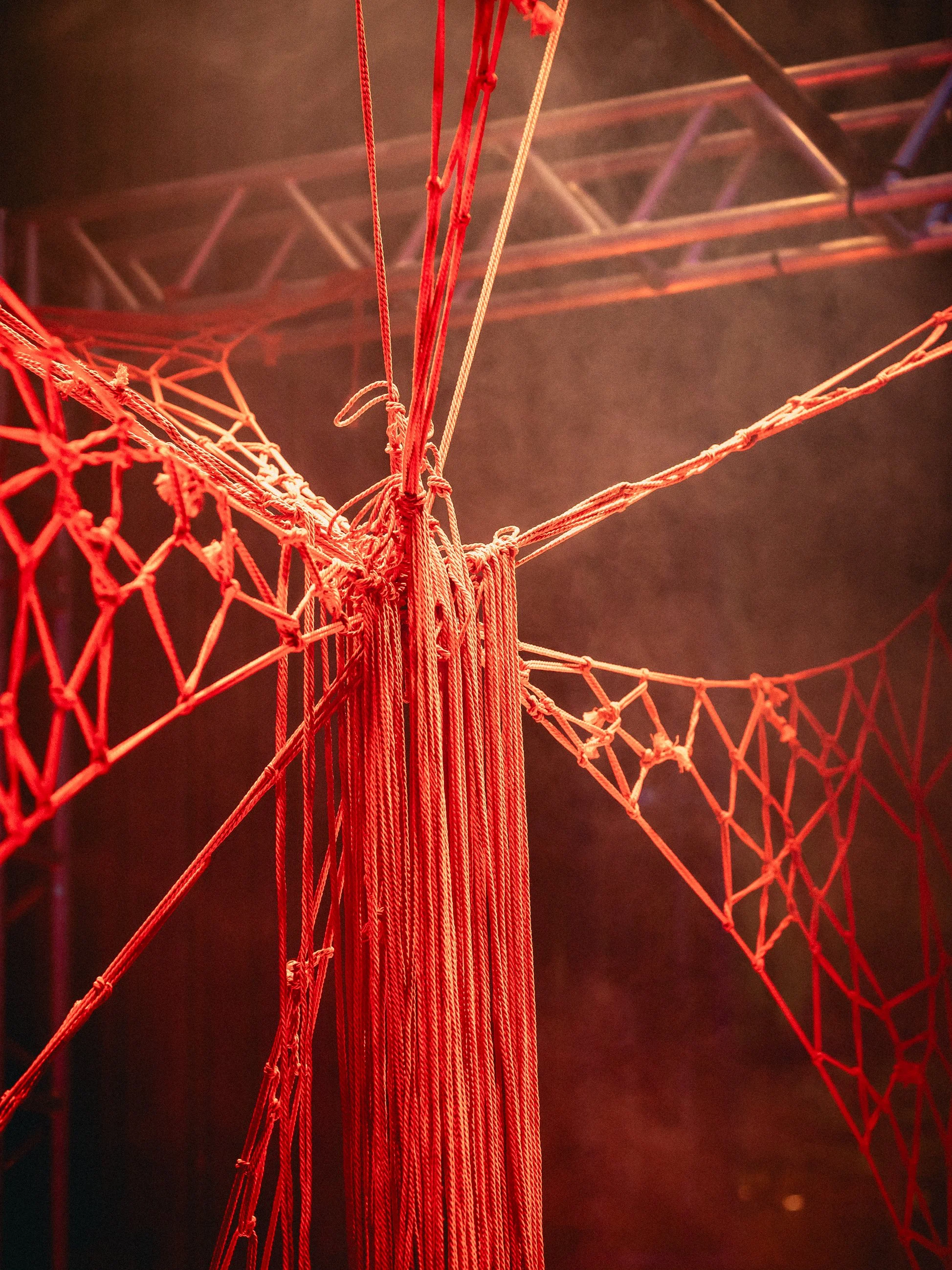 Red ropes and nets tangled around a central pole with some metal support structures in the background, illuminated in red light.