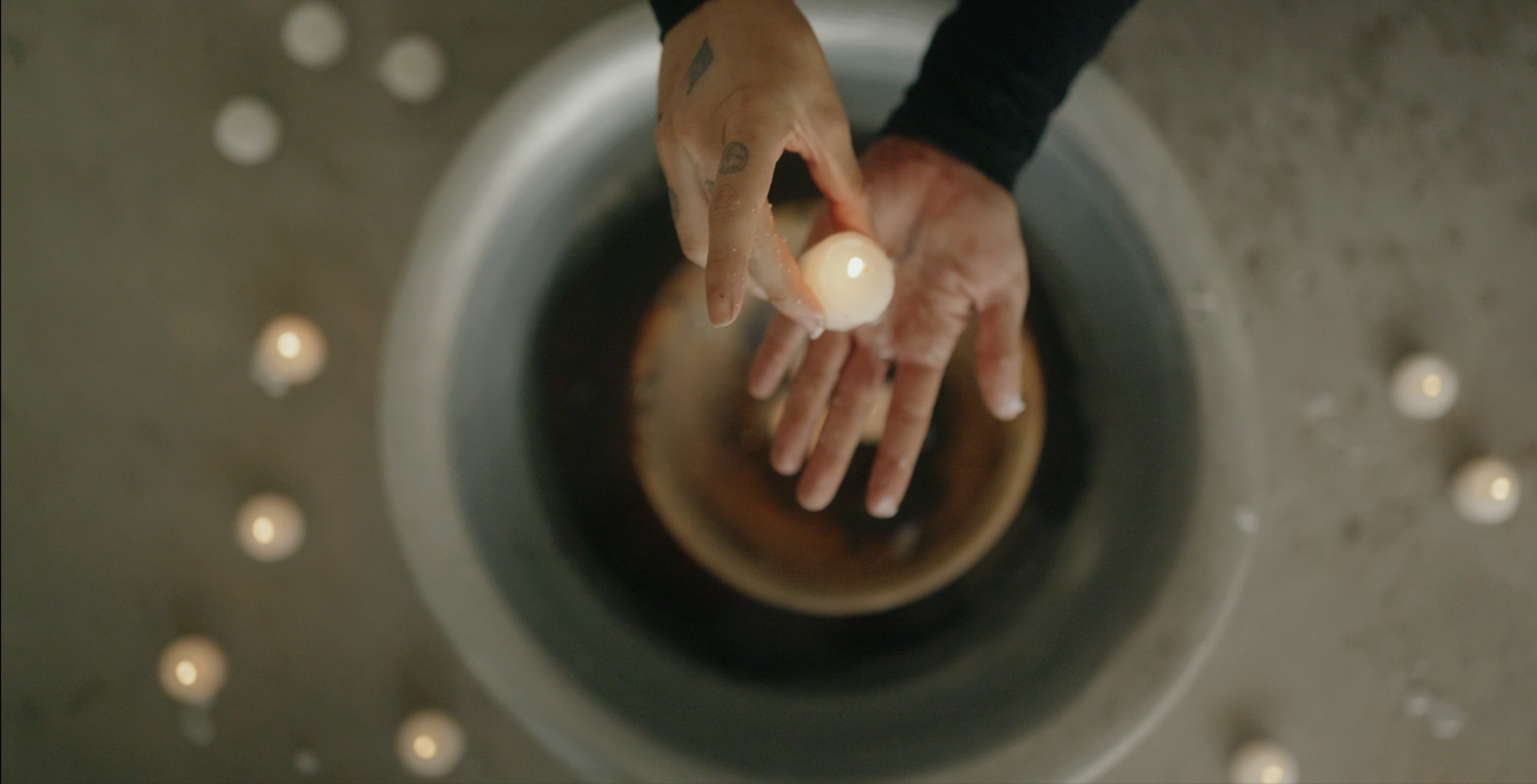 Top-down view of a person's hands holding a small candle over a metal basin, surrounded by small lit candles on a gray surface.
