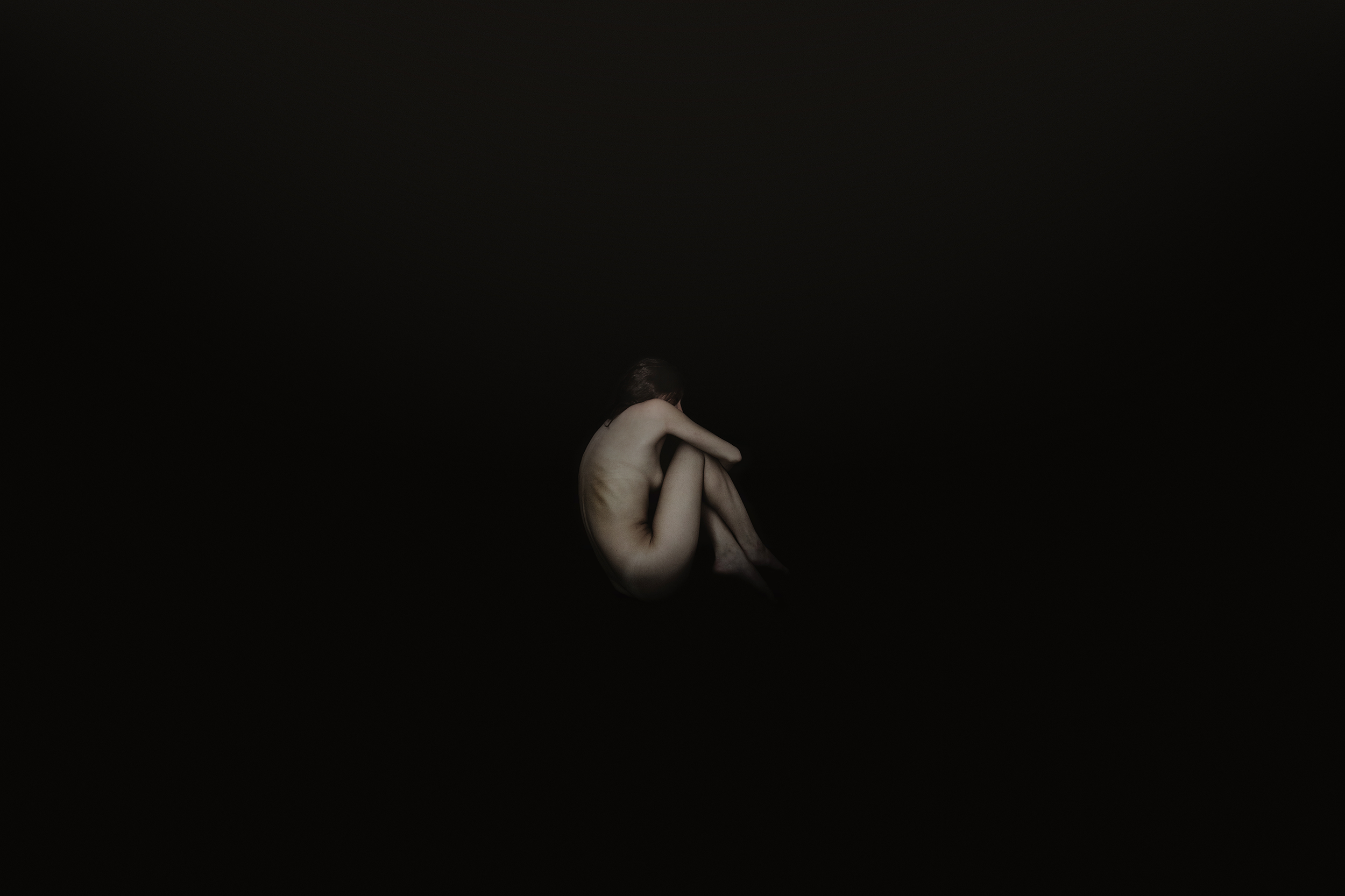 A nude woman sits with knees pulled to chest, head resting on her arm, in a dark, minimalistic setting.