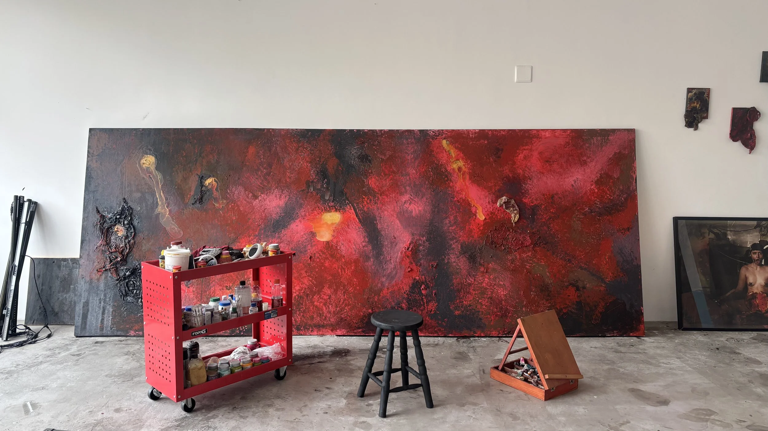 An artist's studio with a large abstract painting in reds, blacks, and oranges leaning against the wall. Various art supplies, including a red rolling cart filled with paints and brushes, a small black stool, a wooden palette box, and framed artwork on the floor and wall, are present.