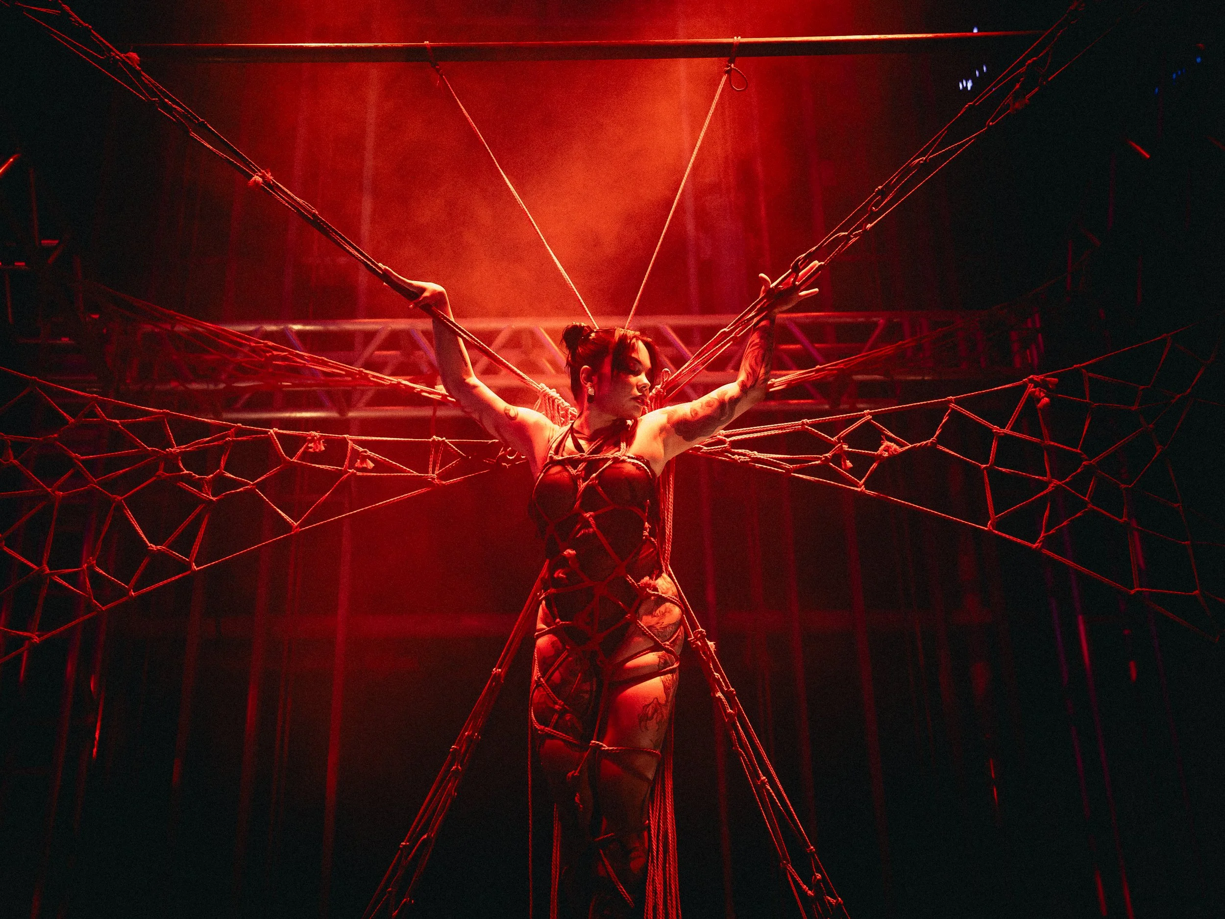 A woman is suspended in the air with ropes, resembling a spider woman silhouette, with red lighting and a stage behind her.
