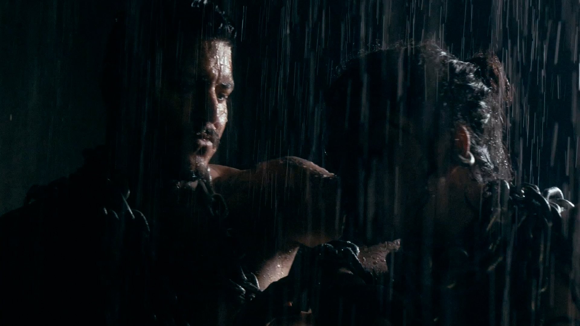 Two individuals are in the rain, one with long hair and earrings is looking at the other, who is partially visible. The scene is dark with water pouring down.