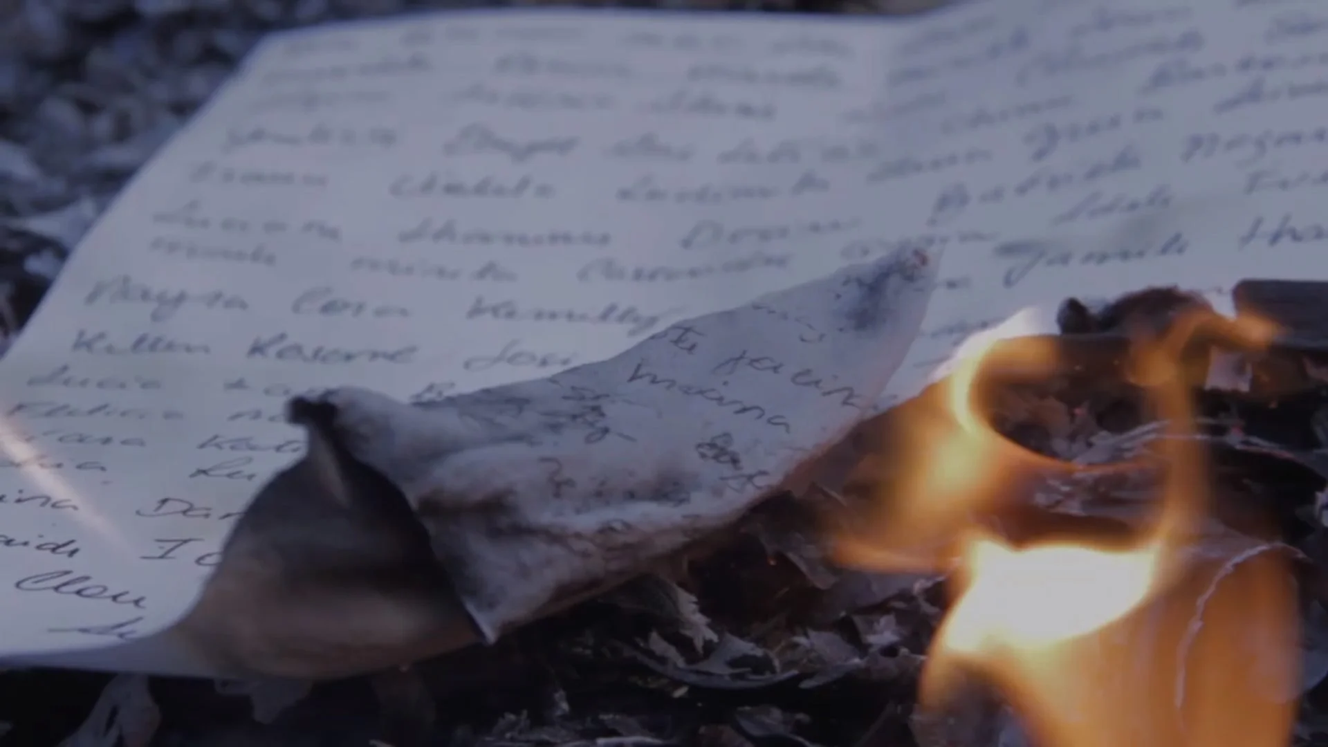 A handwritten letter being set on fire outdoors, with flames visible on the paper.