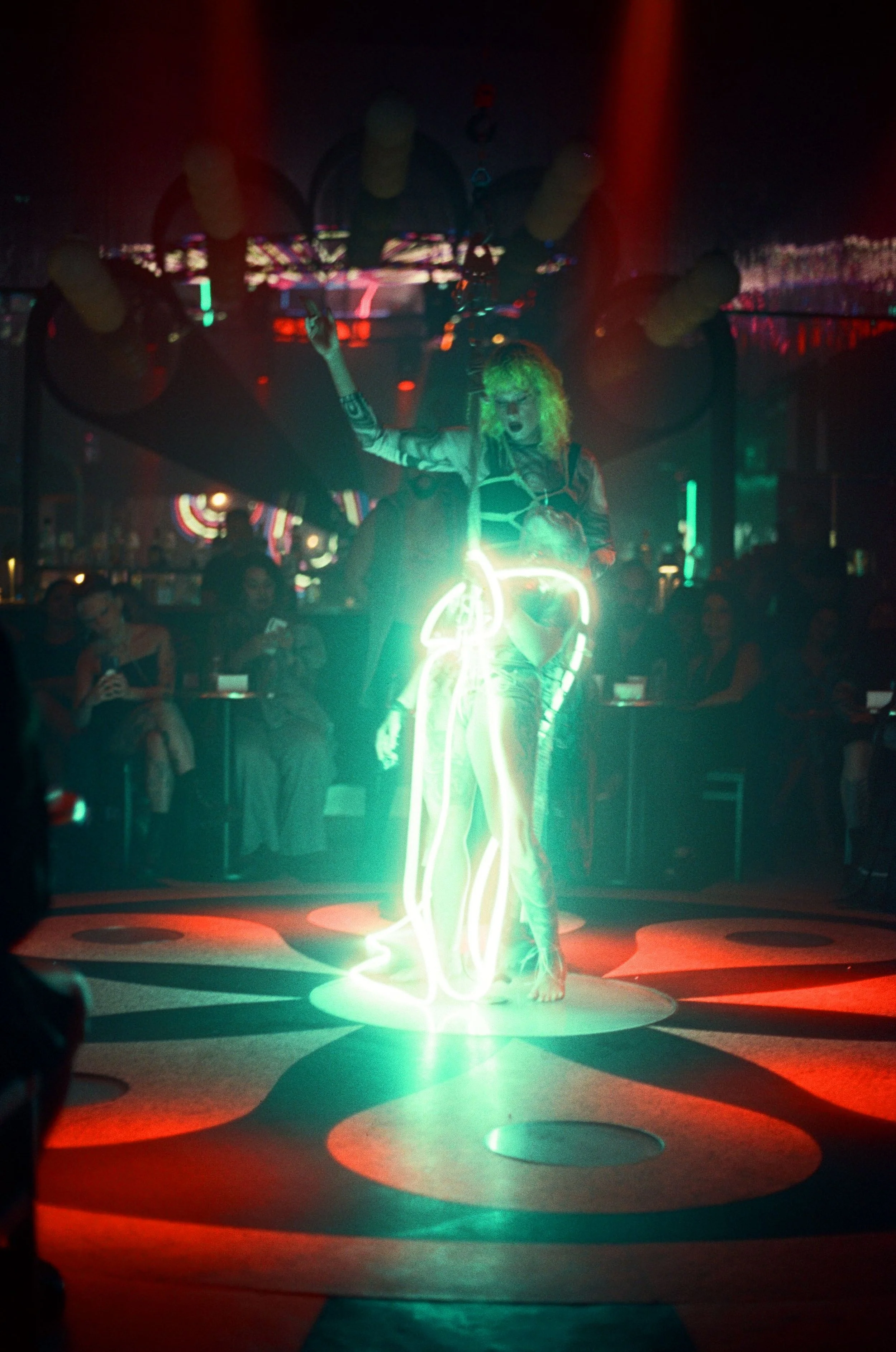 A performer in a nightclub holding a neon light sculpture of a woman, with a crowd watching in the background.