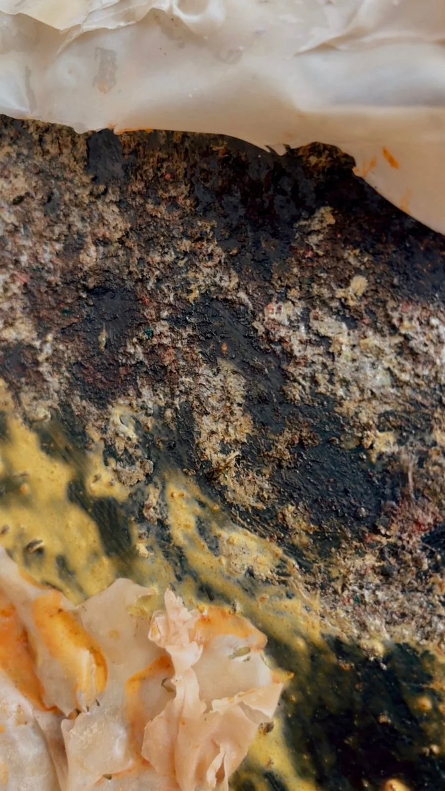 Close-up of a baked dish showing burnt cheese or sauce with darker charred spots and some lighter, creamy parts at the bottom.