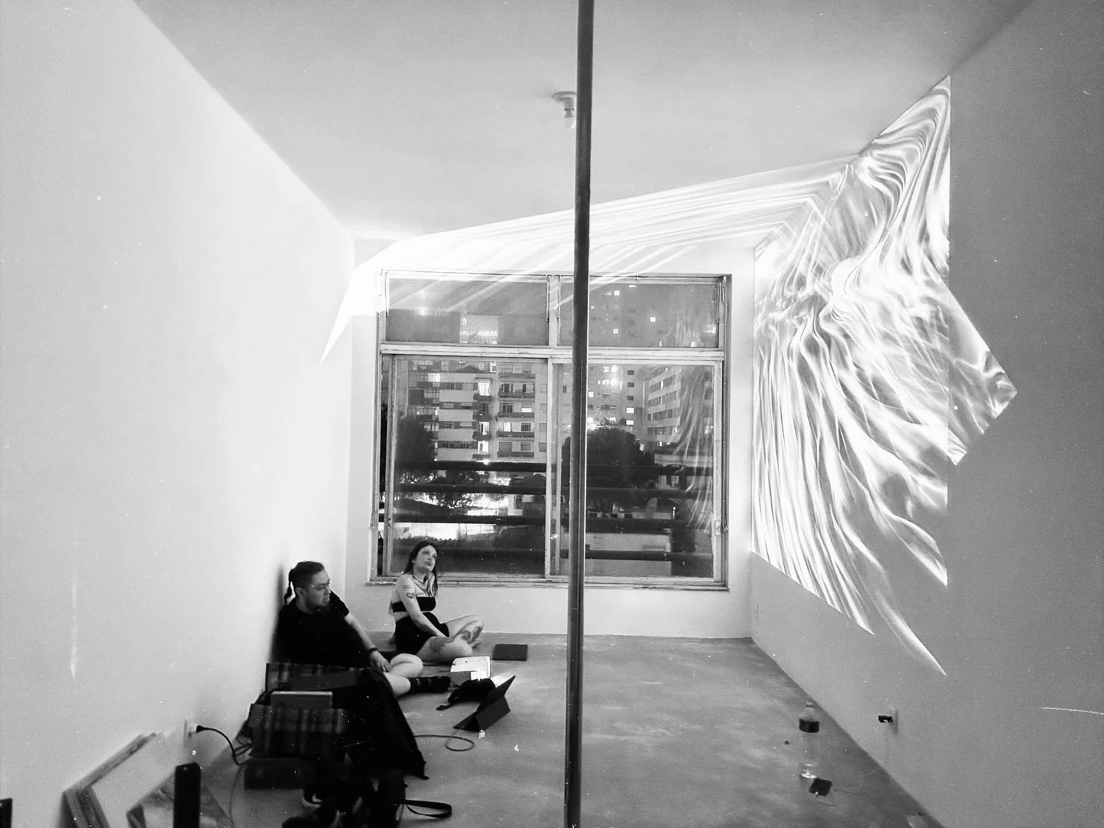 Two people sitting on the floor in an art gallery with a projection of abstract light patterns on the wall behind them.
