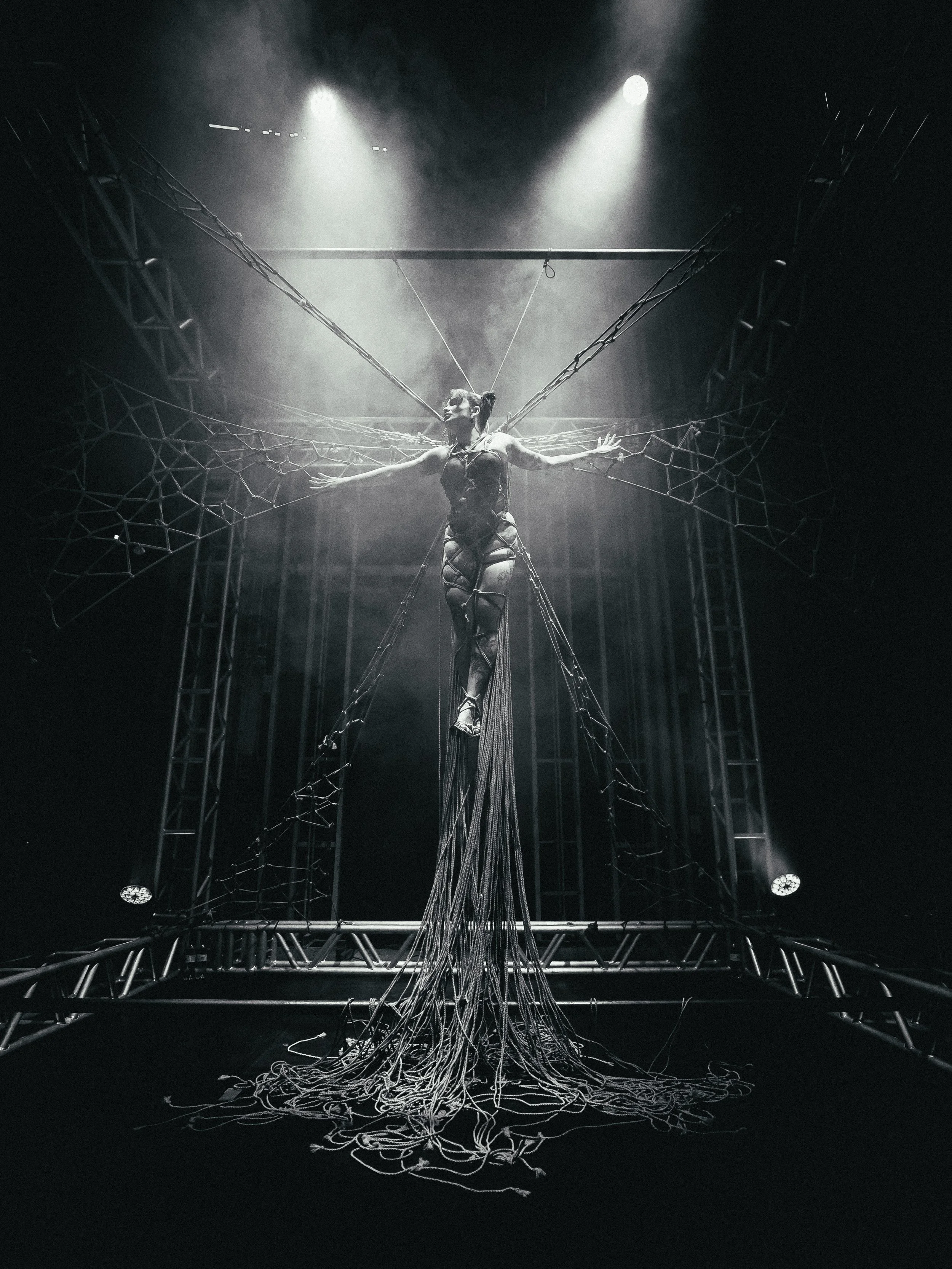 A performer dressed in black is suspended in the air, connected to a web of ropes and metal framework, with stage lights shining down on her in a theatrical production.