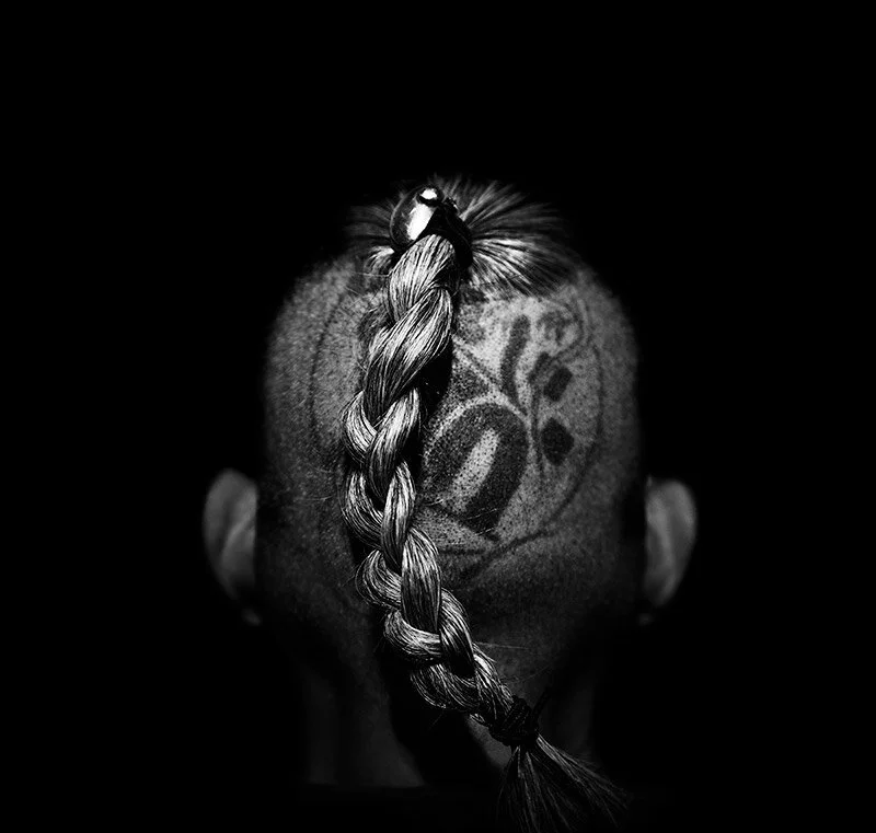 Black and white photo of the back of a person's head with a braid and a tattoo on the scalp.