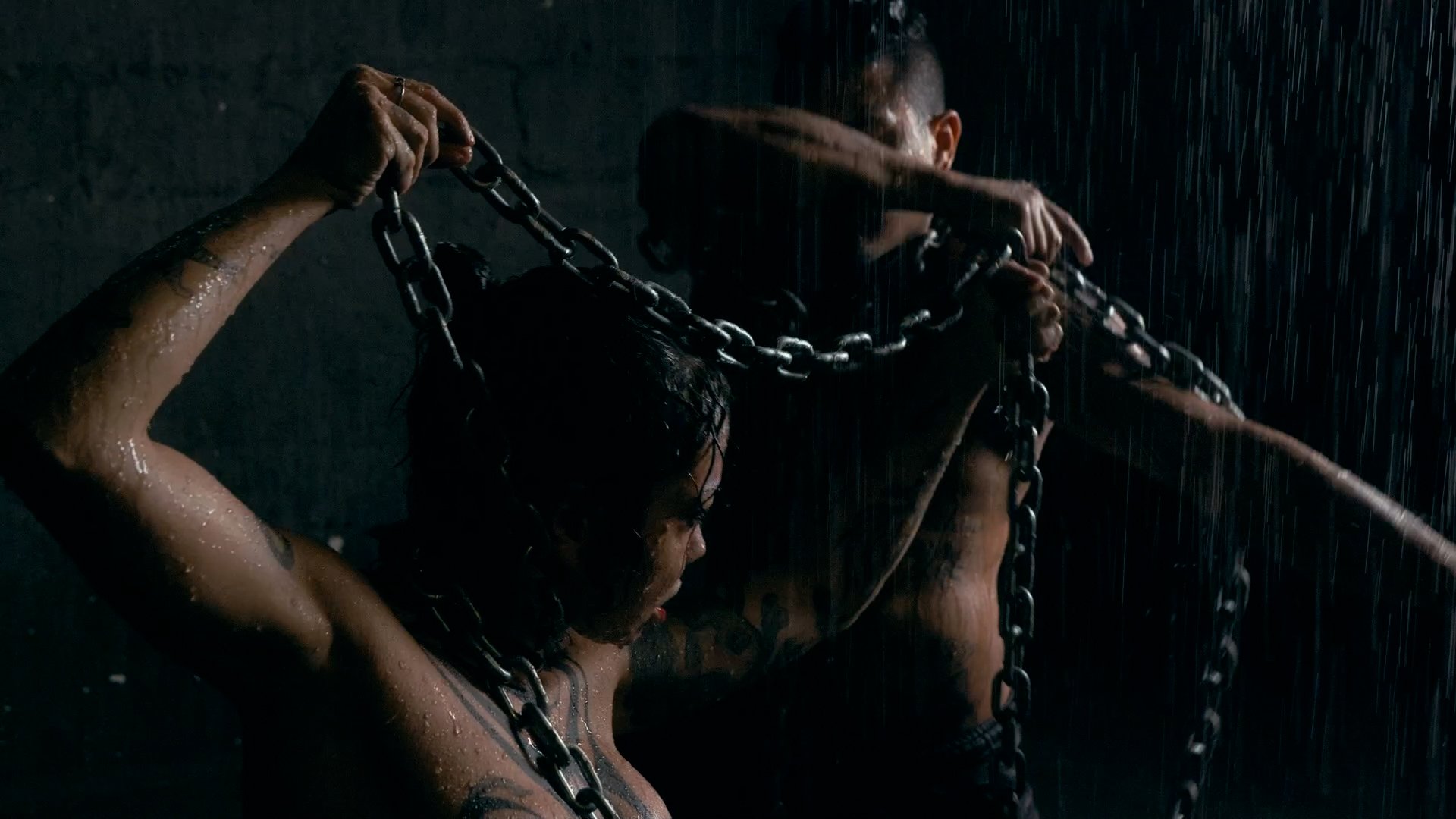 A dark scene showing a woman in the rain holding a chain, with a man behind her also holding chains, both appearing intense.