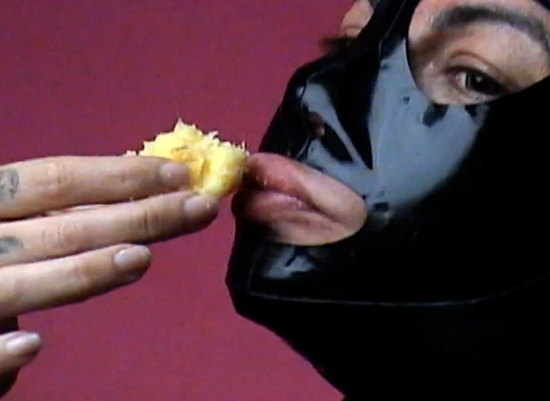 Person in a black jacket and gray shirt with black face mask holding a piece of food, with their face partially visible and an eye visible, against a reddish background.