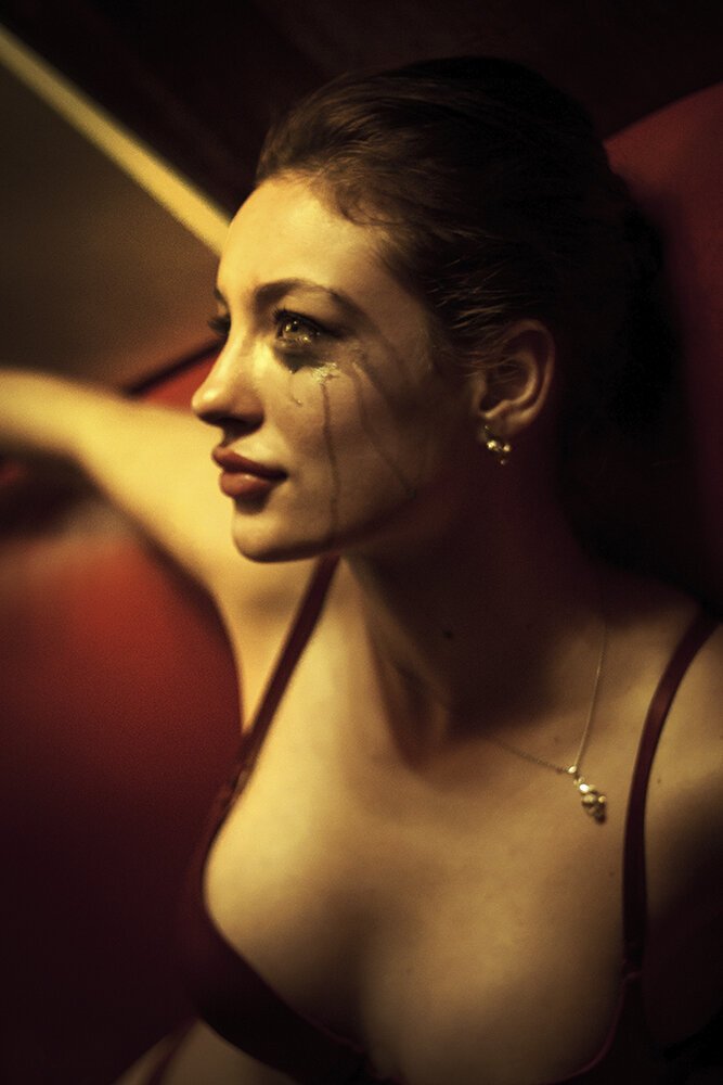 A woman with makeup and tears on her face in a dimly lit setting, wearing a red dress with thin straps and jewelry.