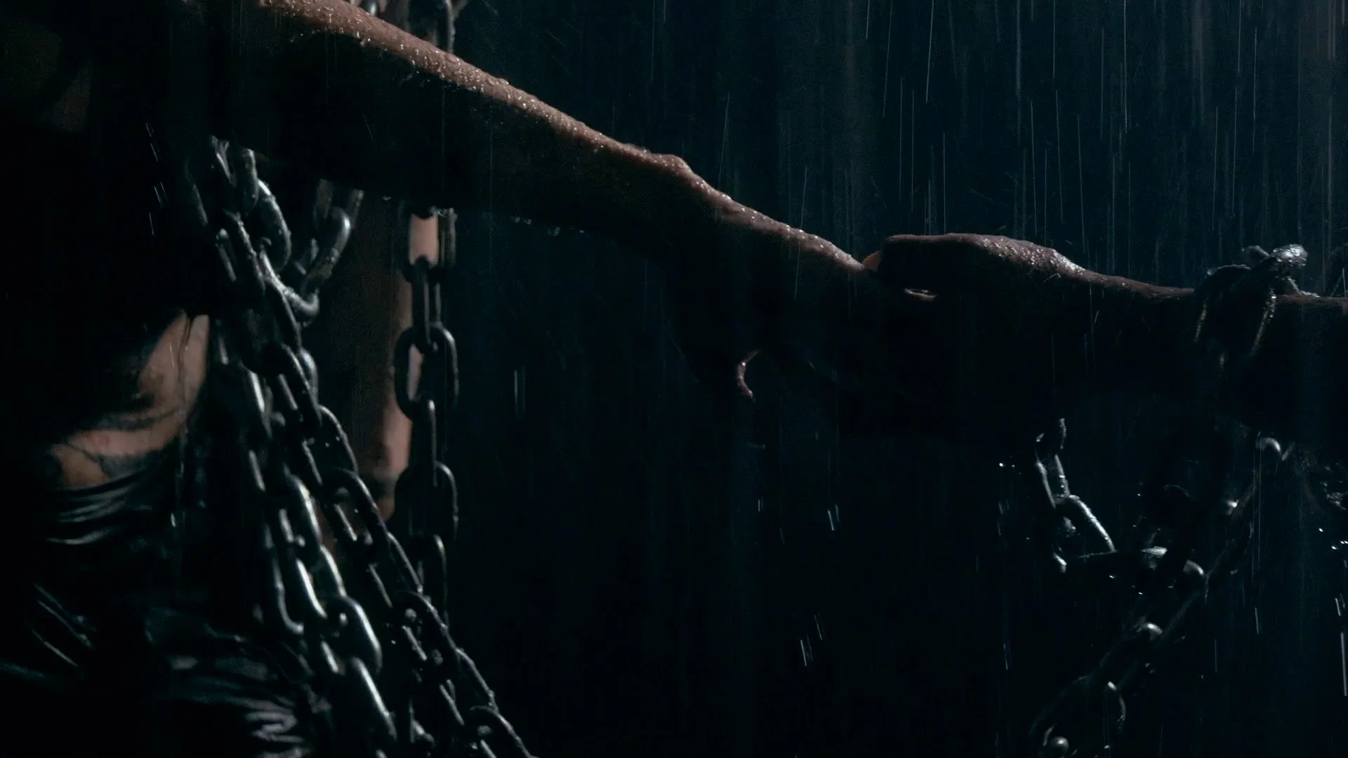 A dark scene with rain falling, showing chains and a fallen tree or branch over a person on the ground.