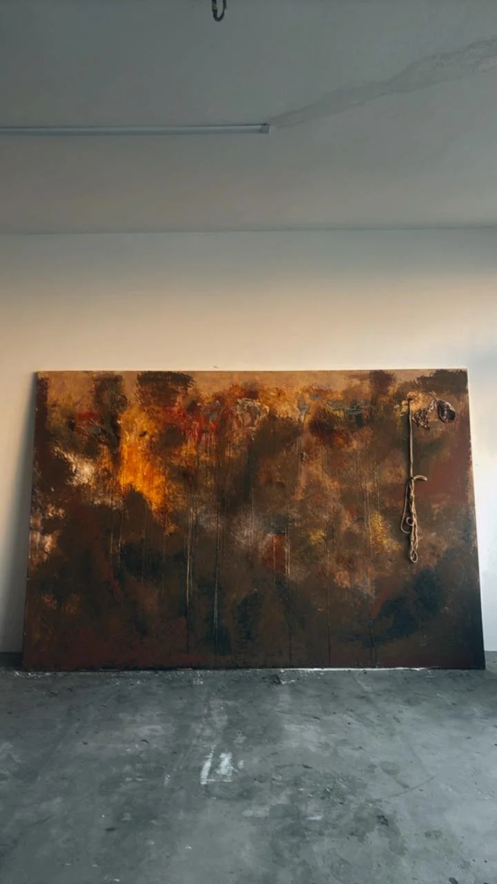 Large abstract painting with dark brown, orange, yellow, and black colors resting against a plain wall in an unfinished room with concrete floor.