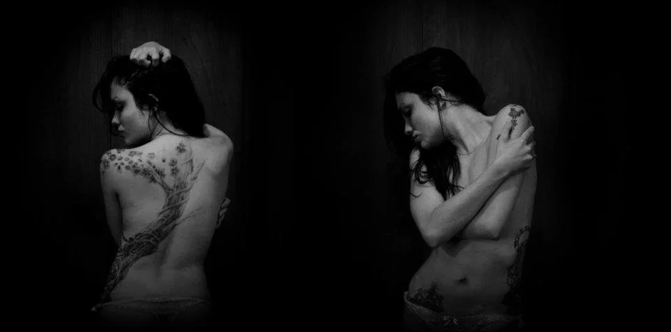 Black and white photo of a woman with tattoos, showing her back and side, with her head bowed and arm over her shoulder.