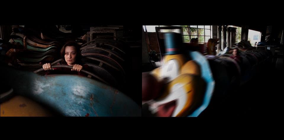 Woman sitting in a carousel with vintage amusement ride horses, looking forward, in a dimly lit indoor space.