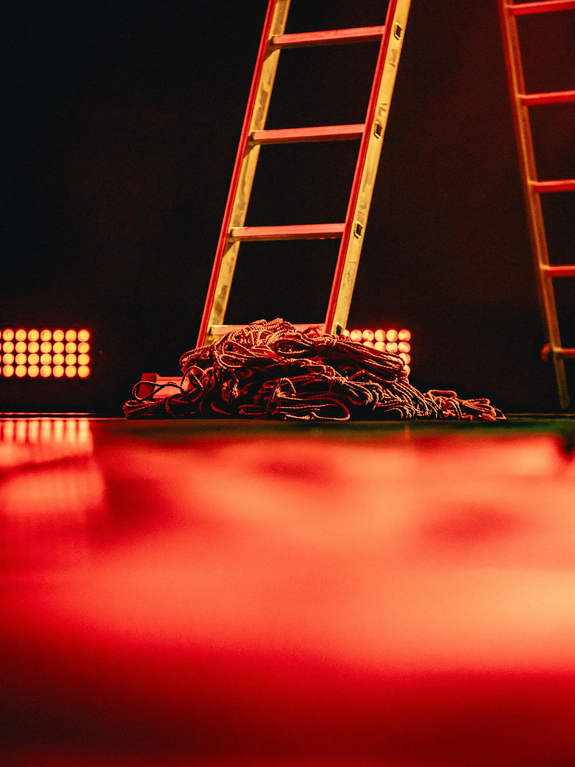 A wooden ladder leaning against a dark background, with a pile of tangled ropes on the floor below, and a red glow from stage lights reflected on the surface.
