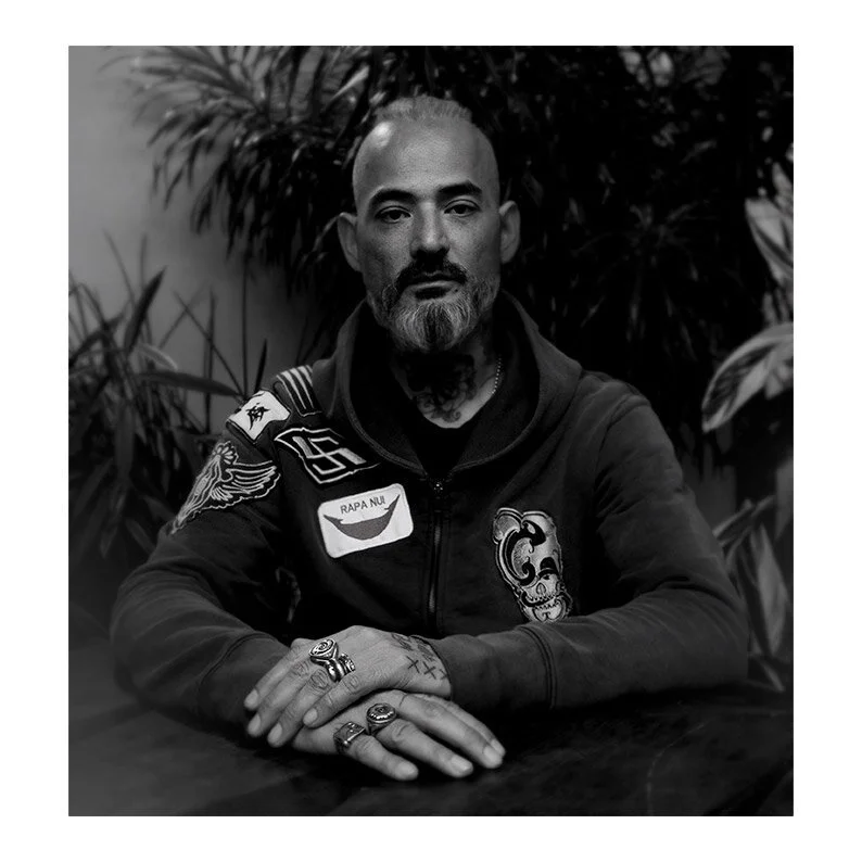Black and white photo of a man with tattoos, facial hair, and a slicked-back hairstyle, sitting at a table with plants in the background, wearing a jacket with patches.