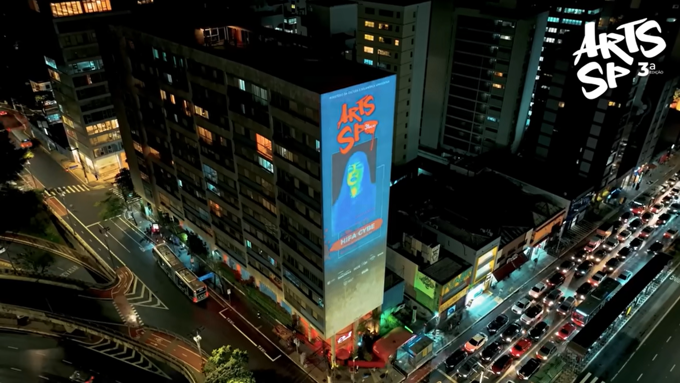 Nighttime city street with a tall building displaying an illuminated billboard for Arts SP 3a. The billboard shows a graphic of a woman with long hair. The street below is busy with traffic, including buses and cars, with some streetlights and buildi