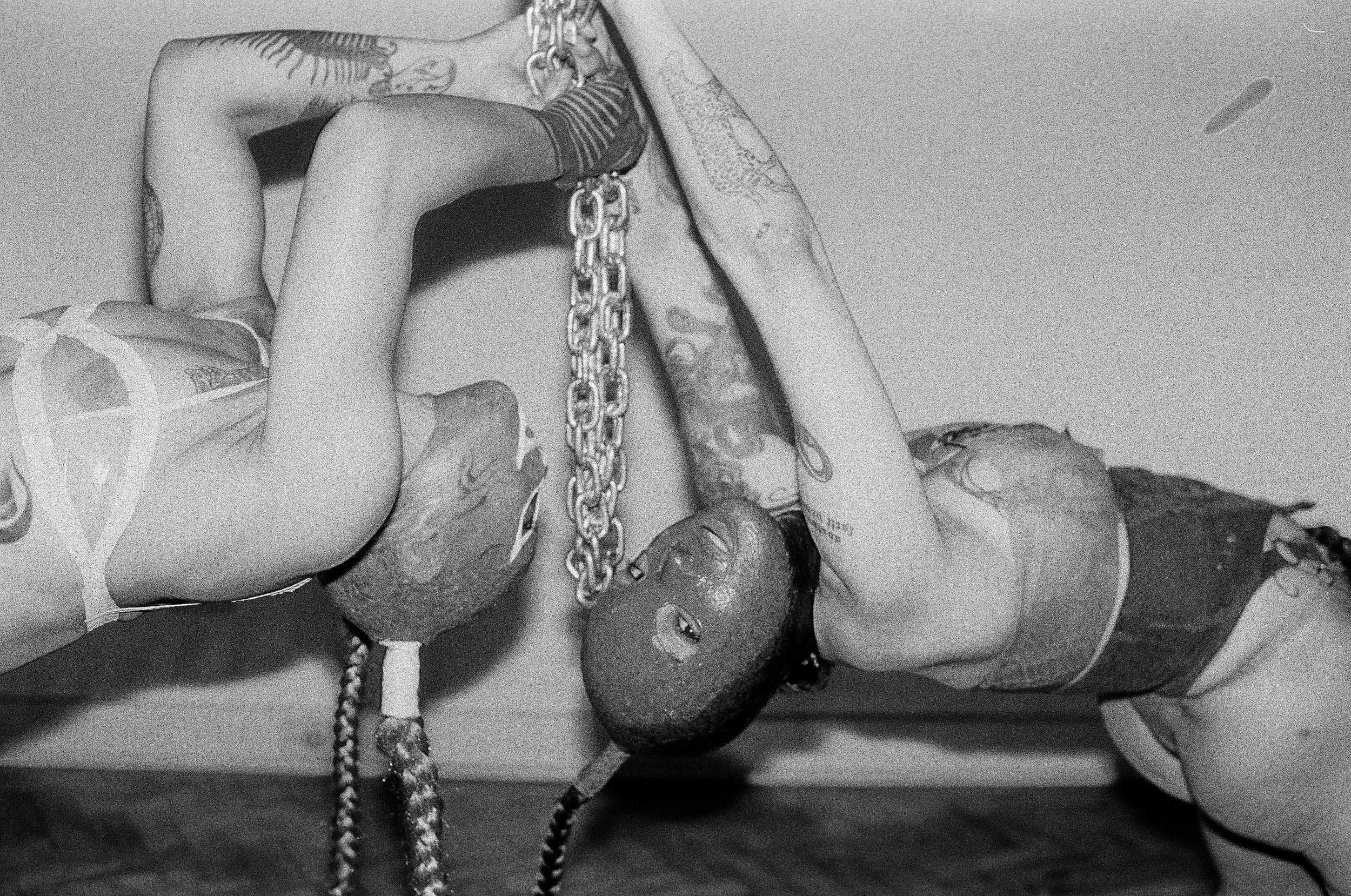 A woman with tattoos lying down and holding a chain, while a man with tattoos and dreadlocks also lies on the floor, their faces close together.