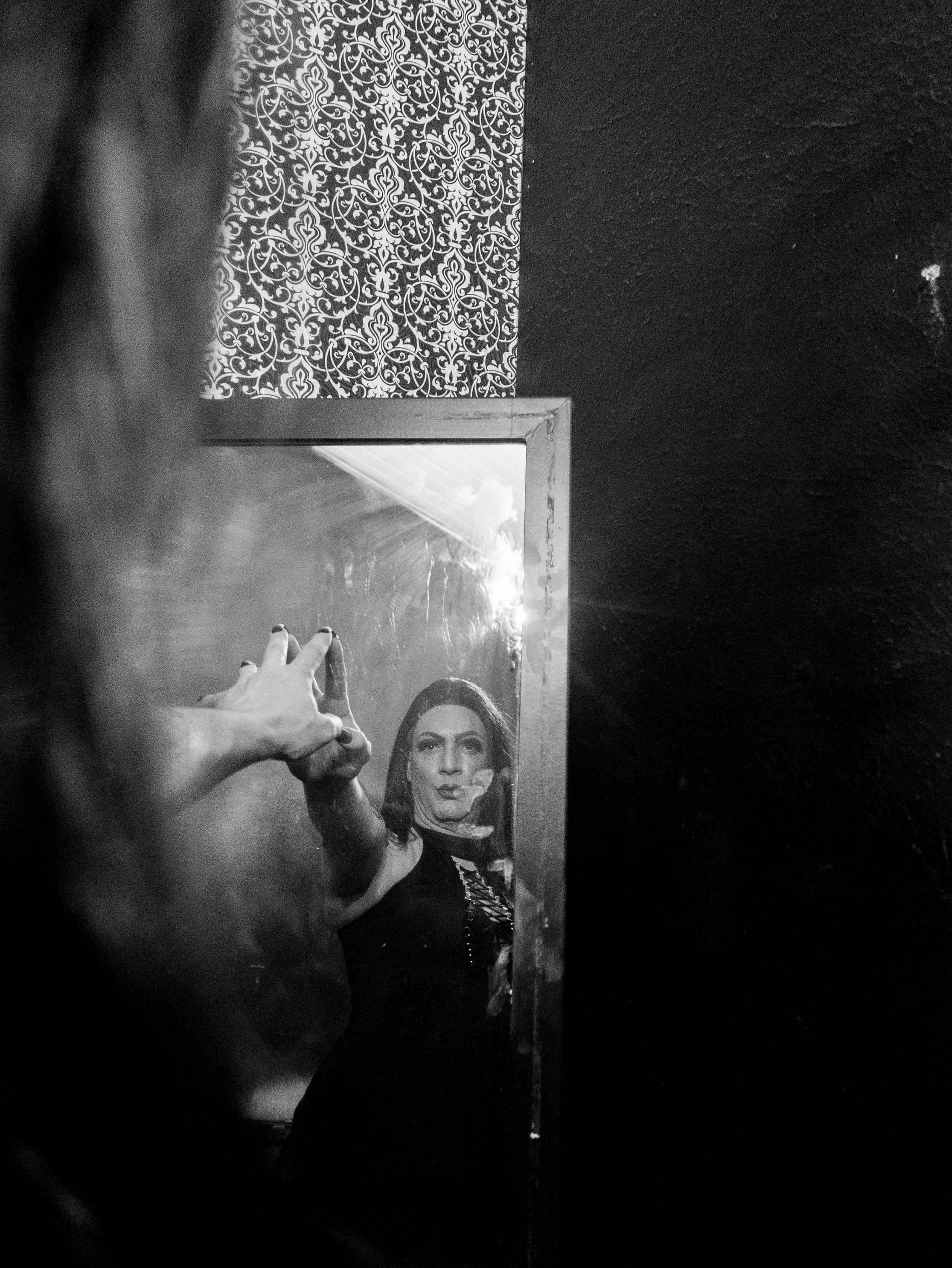 A black and white photograph of a woman taking a selfie in a mirror, with her hand extended and finger pointing at the camera. The woman has dark hair and makeup, and is wearing dark clothing with a necklace. The mirror reflects her face and upper bo
